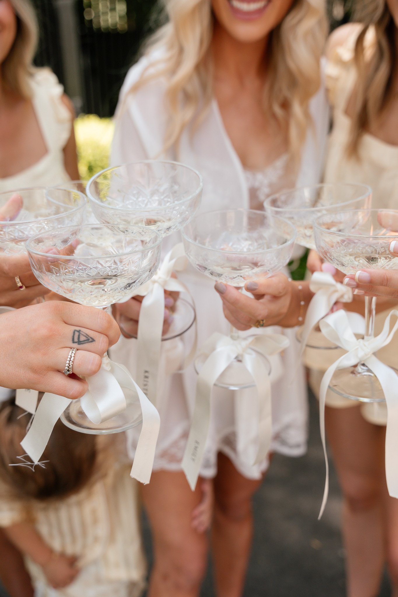 Women in white dresses holding glasses with white ribbons tied around the stems, celebrating at a wedding or party.