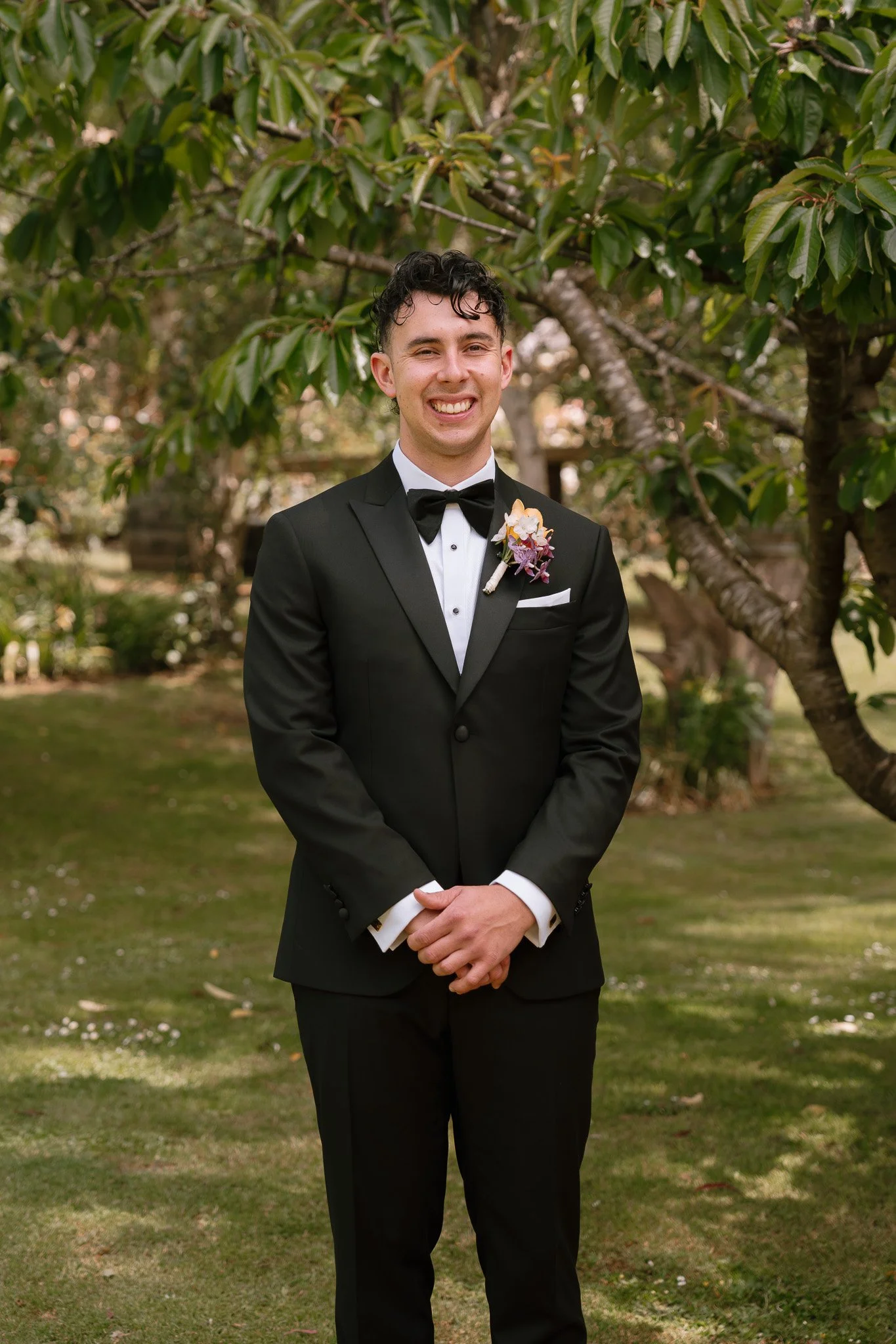 A man dressed in a black tuxedo with a bow tie and a boutonnière, smiling and standing outdoors near a tree.
