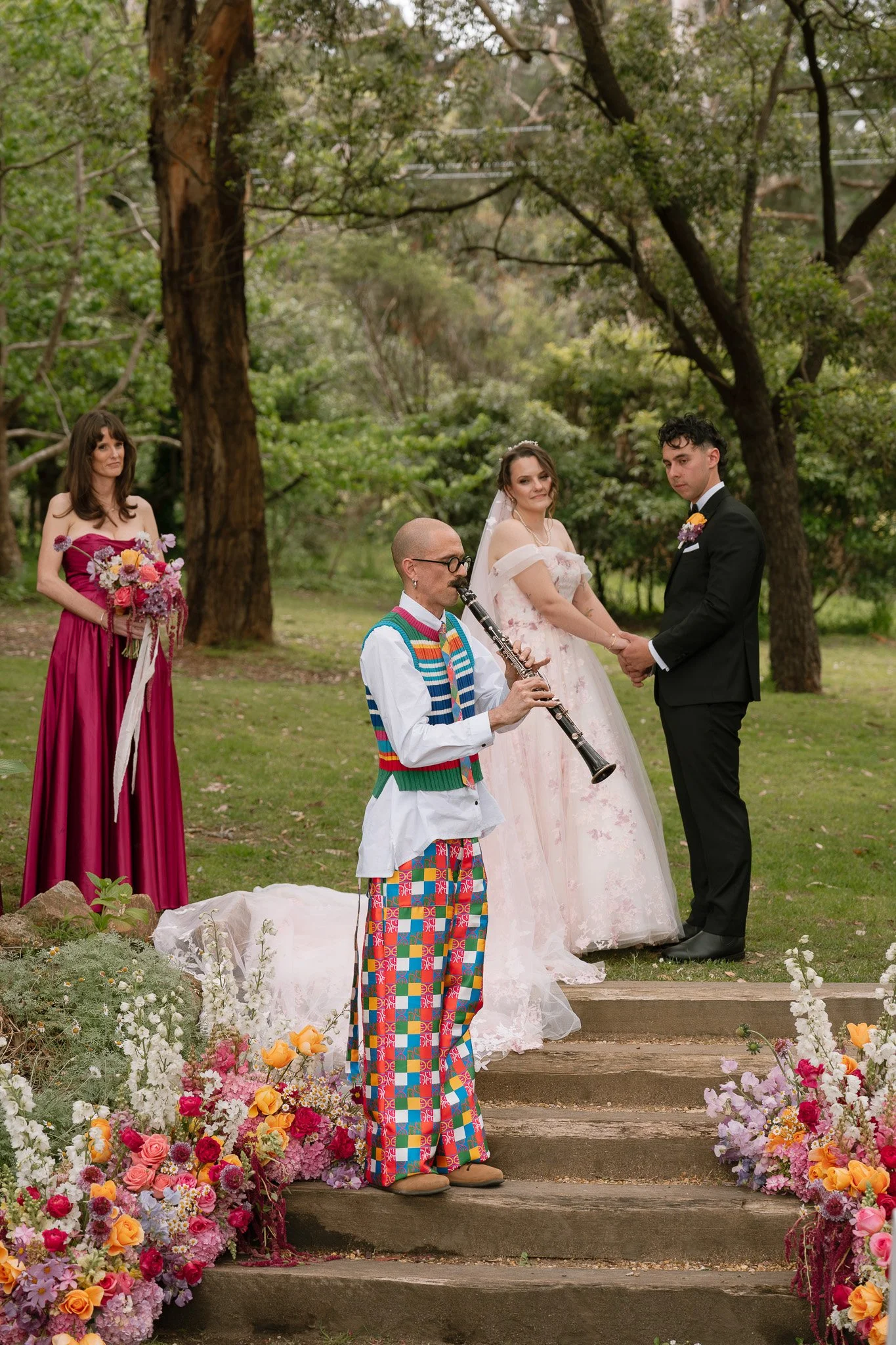 A wedding ceremony taking place outdoors in a wooded area. The bride and groom are holding hands, with the bride wearing a white gown and the groom in a black suit. A person dressed in colorful, patterned clothing is playing a clarinet nearby. A woma