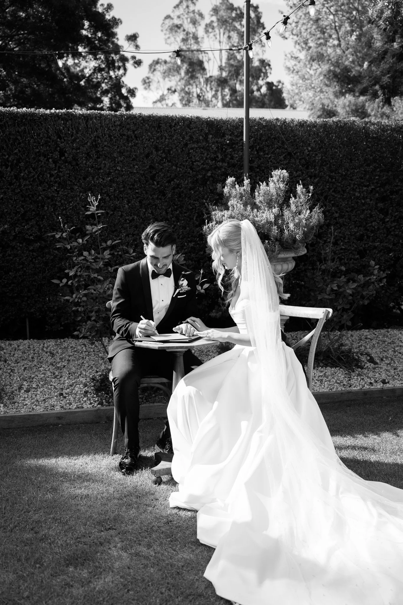 Black and white photo of a bride and groom signing a marriage register outdoors, with the bride in a wedding gown and veil, and the groom in a tuxedo, near a hedge and decorative plants.