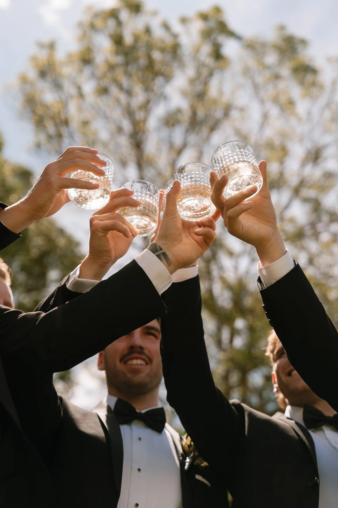 A group of men in tuxedos toasting with glasses of clear liquid outdoors during daytime.