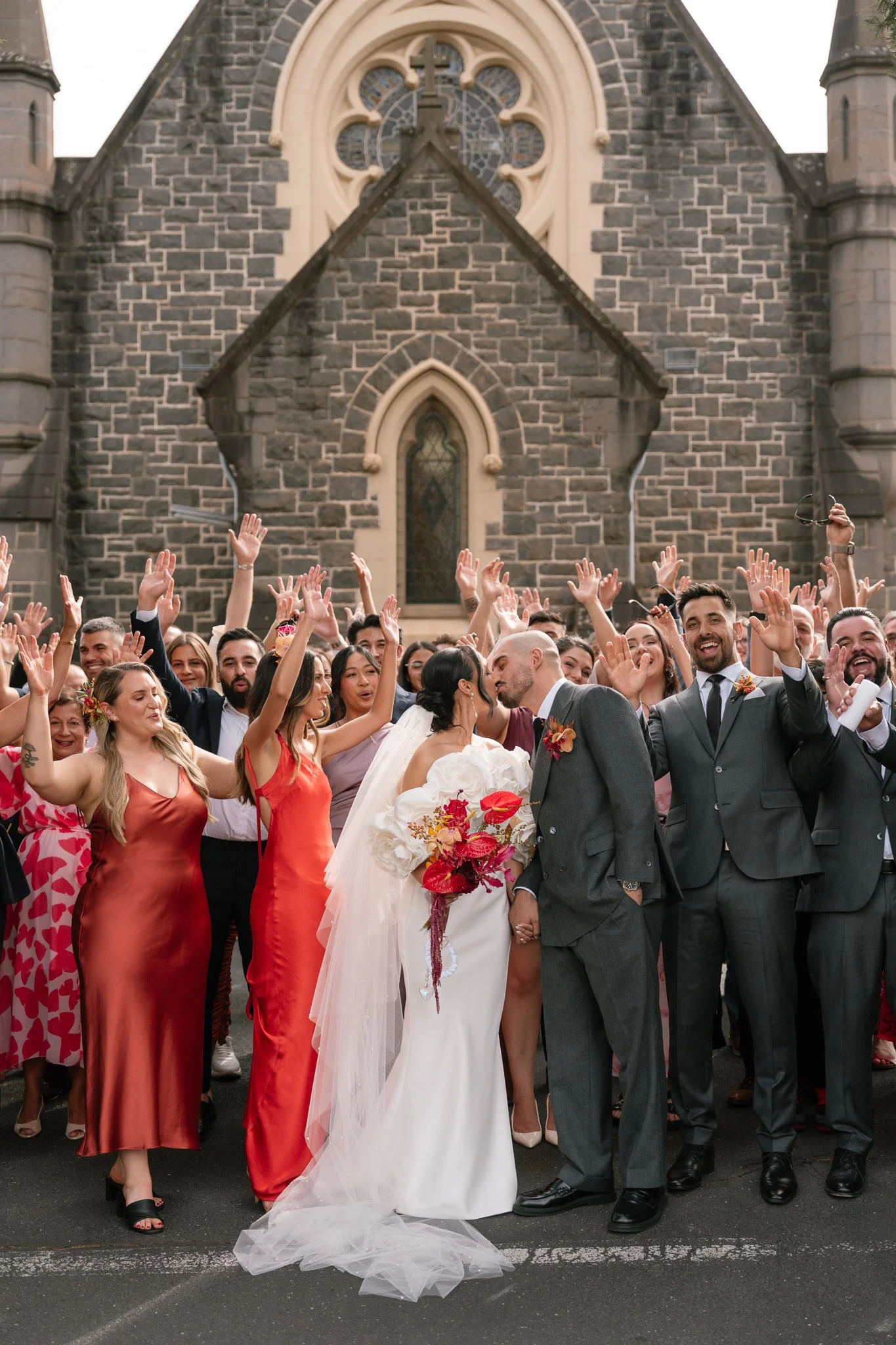 A wedding celebration in front of a church with a large group of guests. The bride and groom are kissing, holding hands, surrounded by guests in colorful attire, and the church's stone facade is visible in the background.