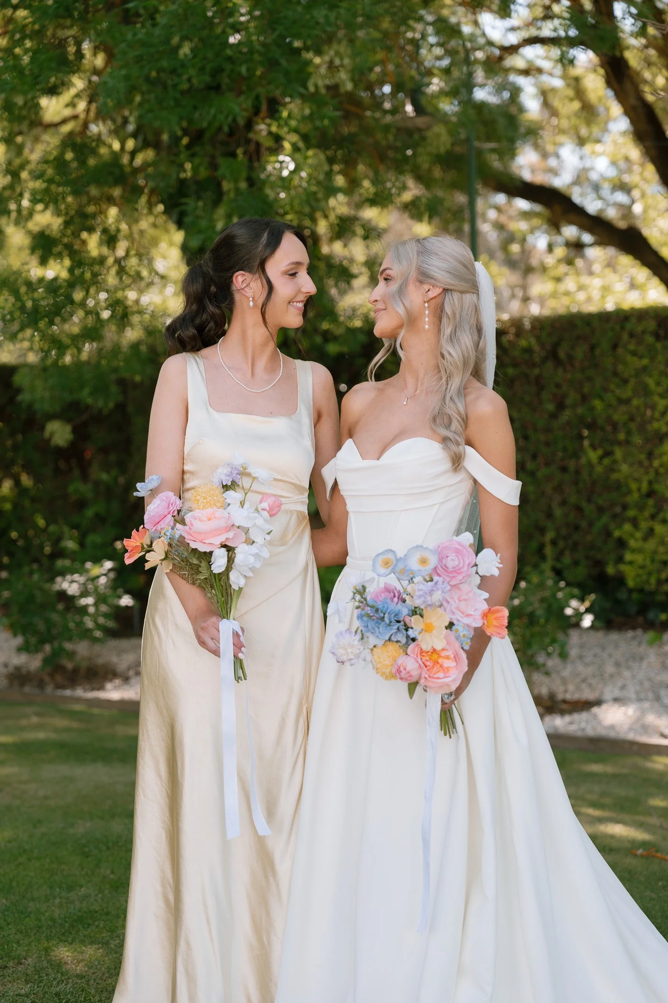 Two women in wedding dresses holding bouquets looking at each other in a garden.