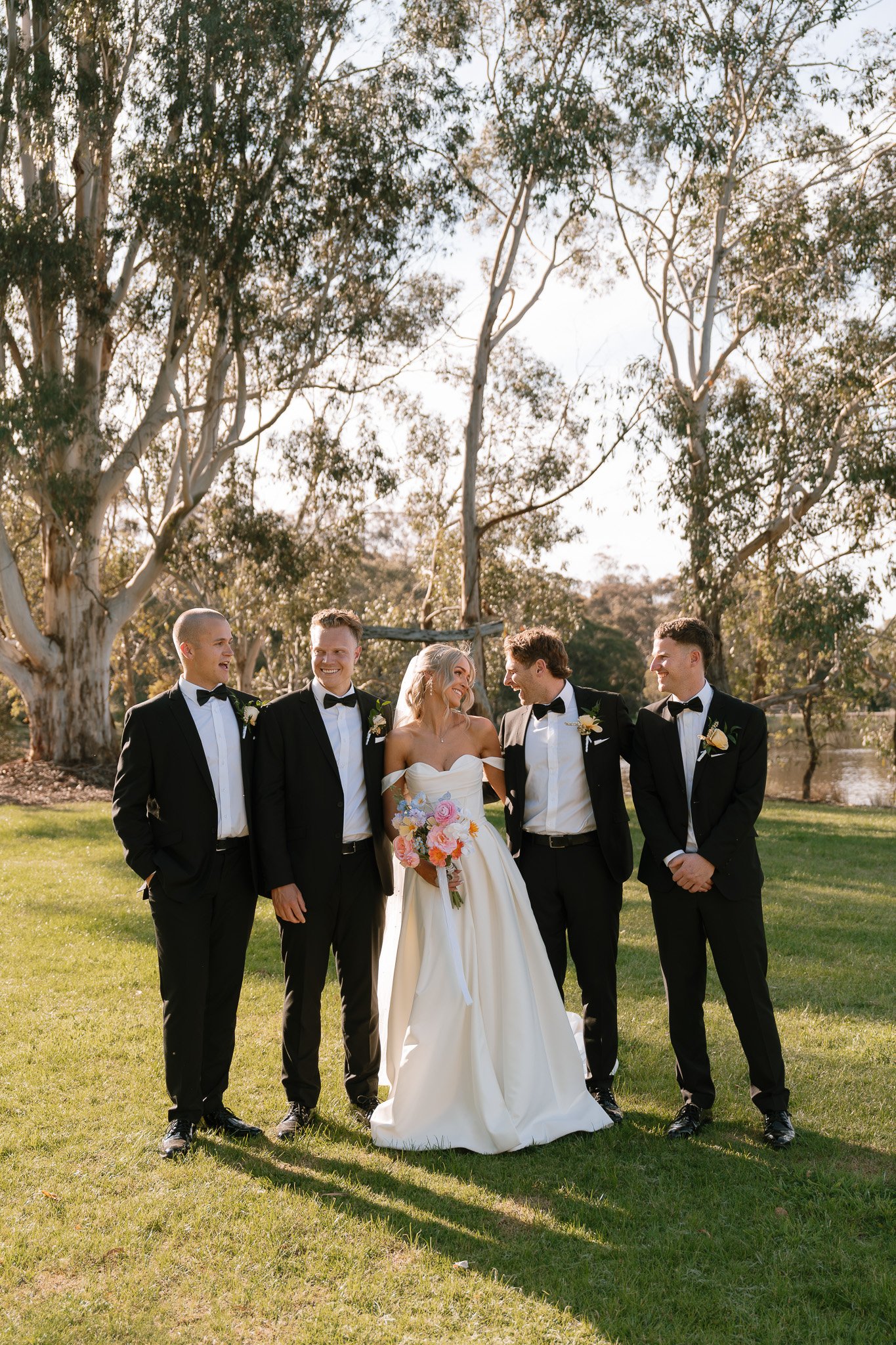 A bride in a white wedding gown holding a bouquet of flowers and five groomsmen in black tuxedos standing outdoors with trees and a pond in the background.