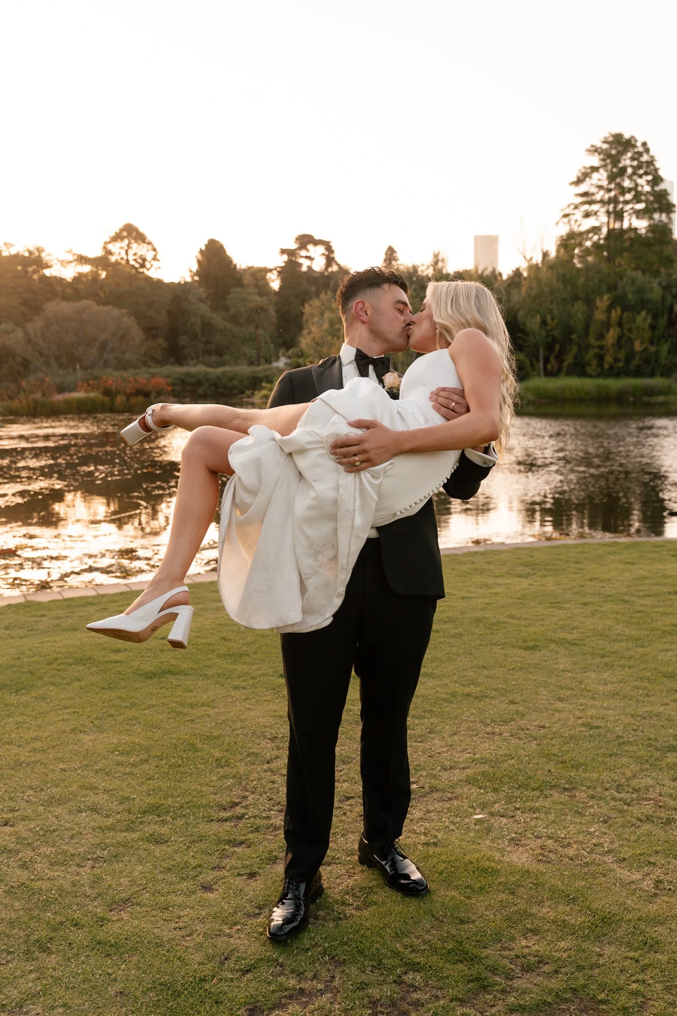 A groom in a tuxedo holding a bride in a white wedding dress and high heels, kissing near a lake during sunset.