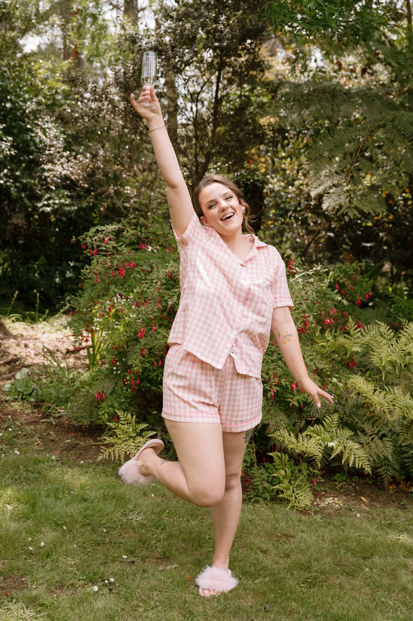 A young woman in pink checkered pajamas celebrating outdoors, holding a glass of champagne and smiling.