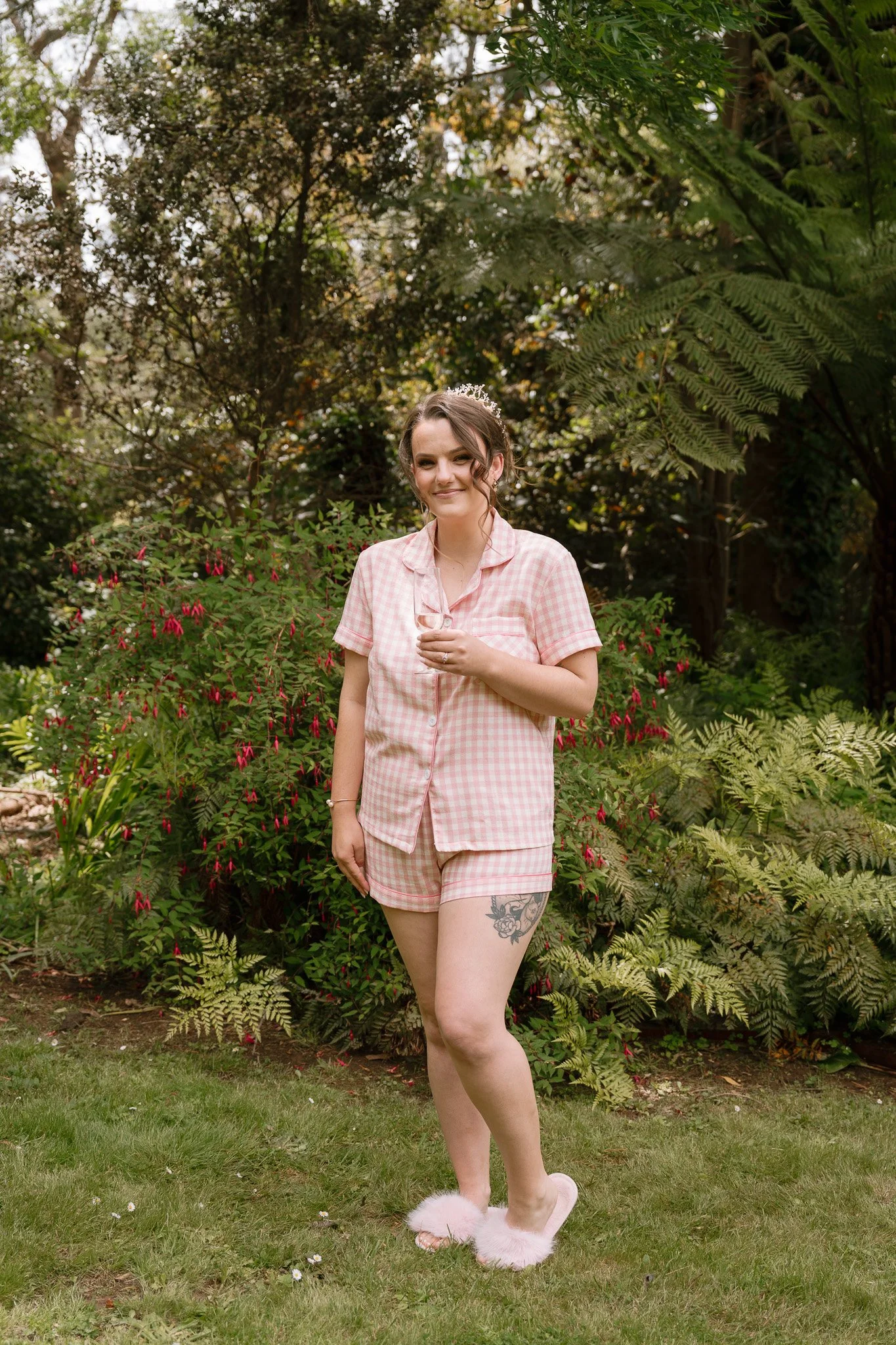 A young woman in pink checkered pajamas and fluffy slippers standing outdoors in a garden with green foliage and pink flowers, holding a glass of wine and smiling.