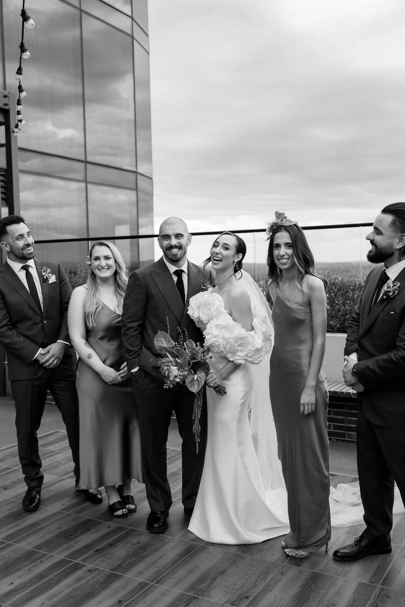Black and white photo of a wedding party on a rooftop, featuring a bride in a white dress holding a bouquet, surrounded by five wedding guests in formal attire, with a modern building and cloudy sky in the background.