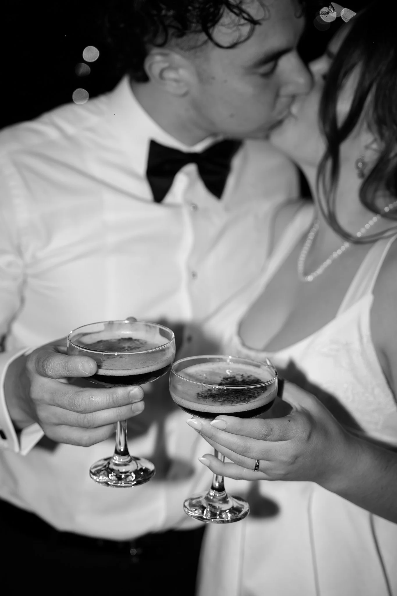 A black and white photo of a couple kissing while holding cocktails in coupe glasses, dressed in formal attire including a tuxedo and a white dress.