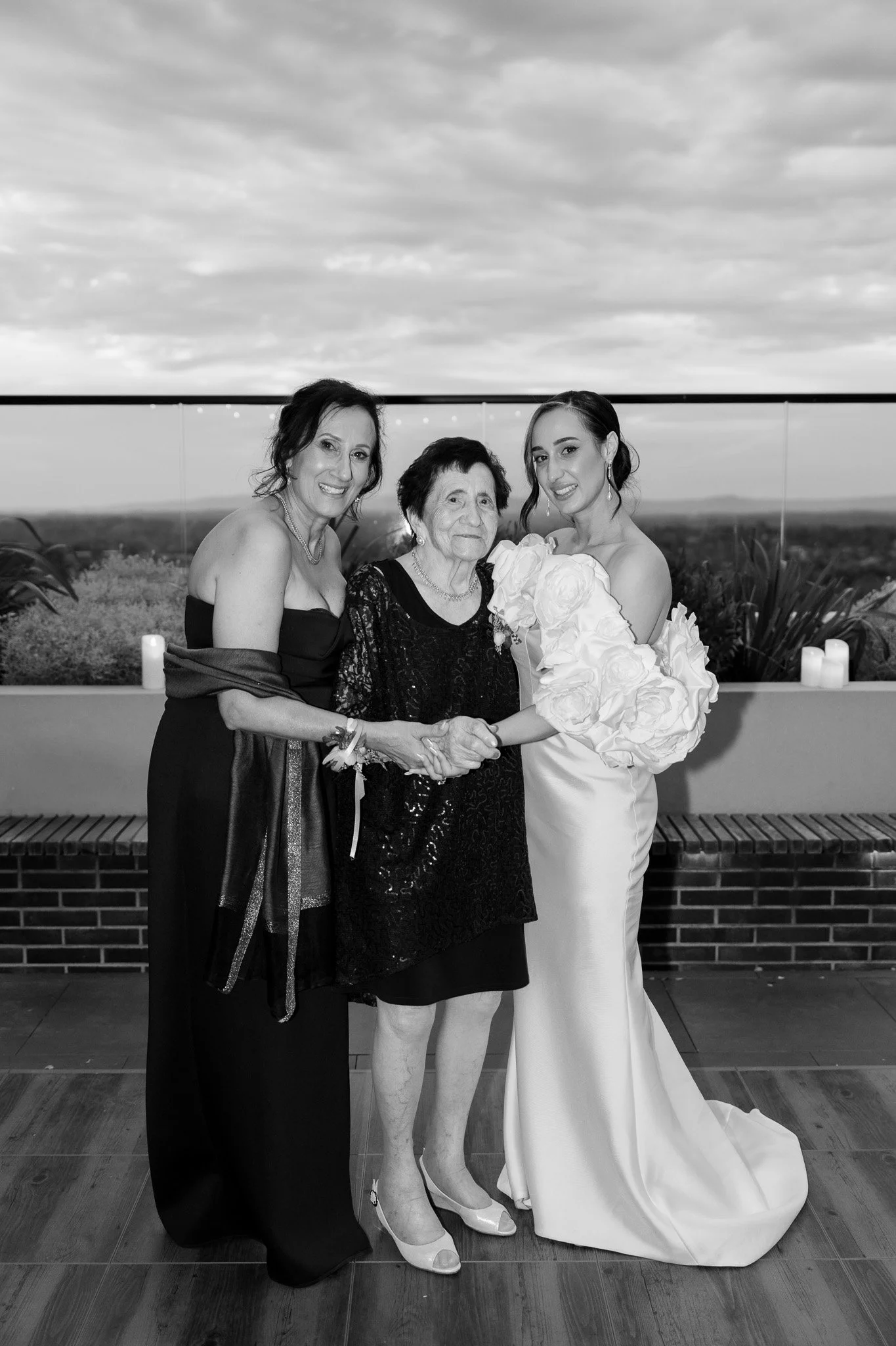 Black and white photo of three women at a celebration, possibly a wedding, standing on a rooftop with a scenic landscape in the background. The woman on the right is wearing a wedding dress and holding a bouquet of large flowers. The woman in the mid
