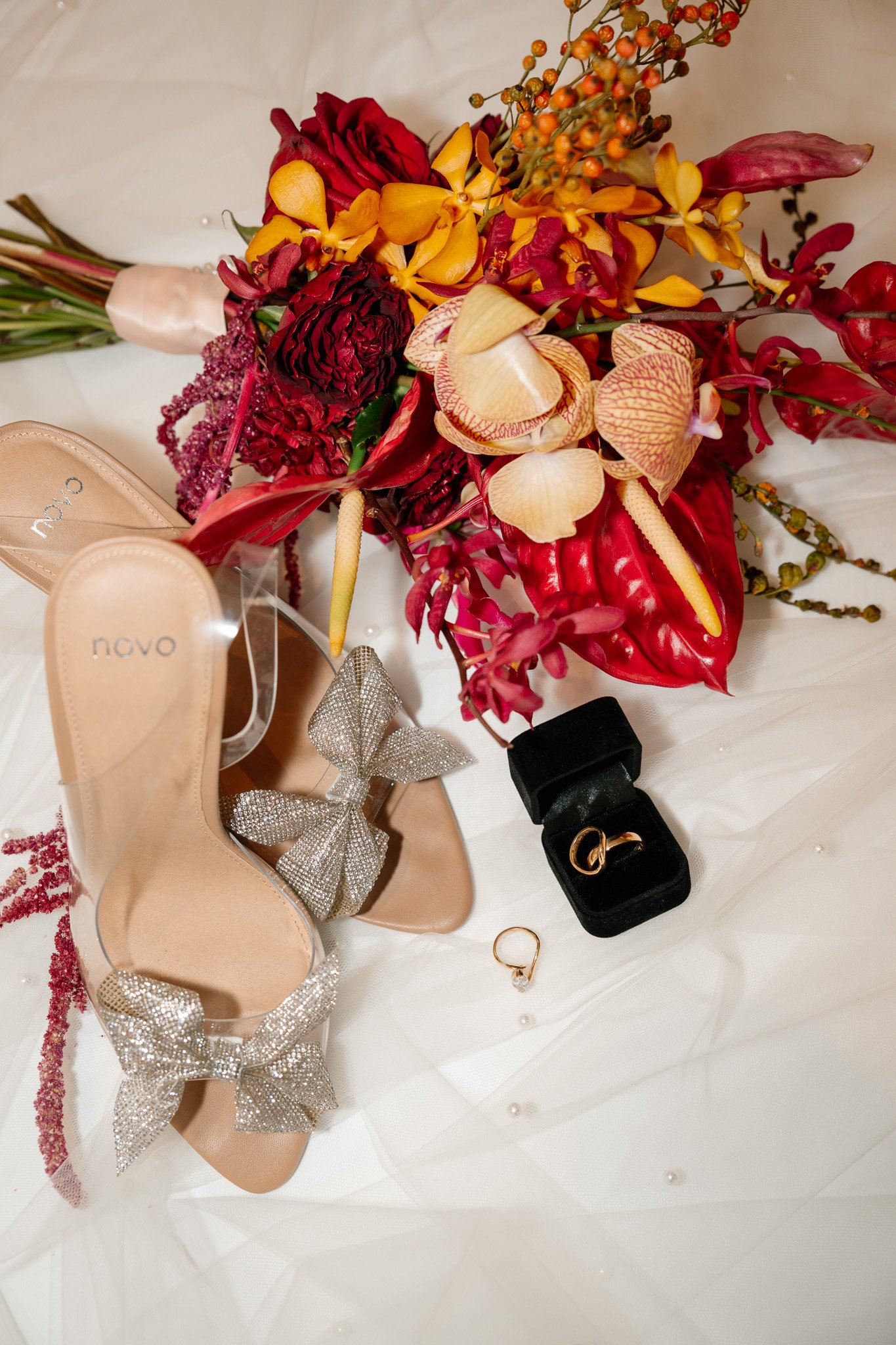 A bouquet of red, yellow, and peach flowers and green foliage, a pair of beige high heel shoes with large sparkling bows, a small black ring box with gold rings inside, and a single gold ring with a gemstone, all arranged on a white surface with scat