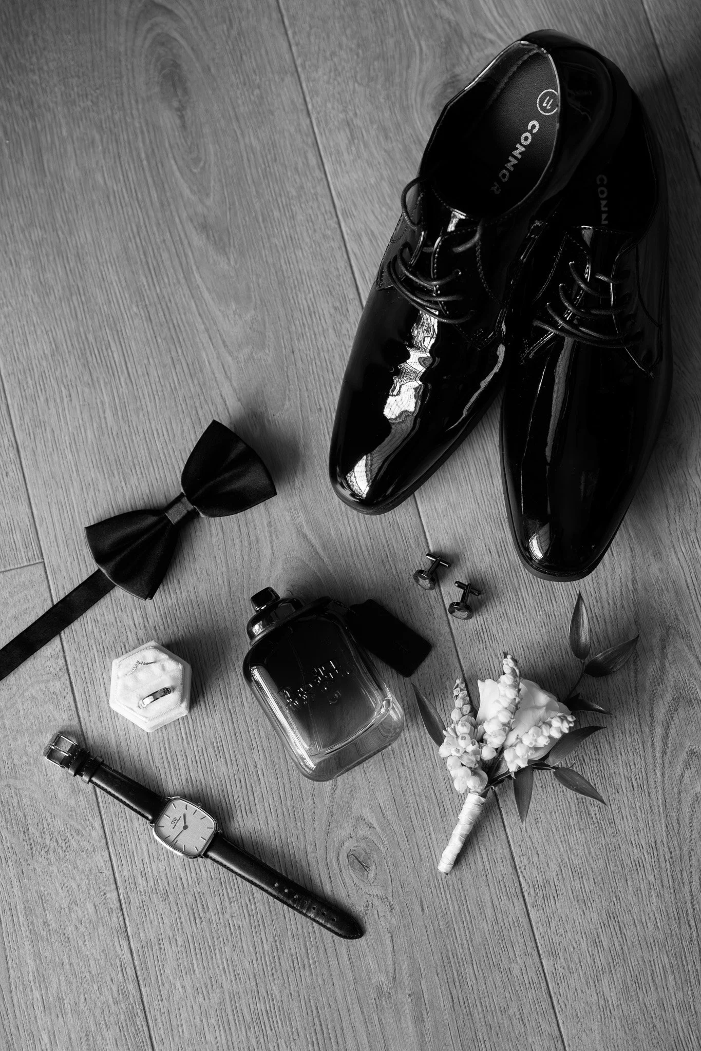 Black dress shoes, a wristwatch, a perfume bottle, a black bow tie, a small flower boutonniere, a ring in a box, a pair of cufflinks, and a watch in a hexagonal case on a wooden floor.