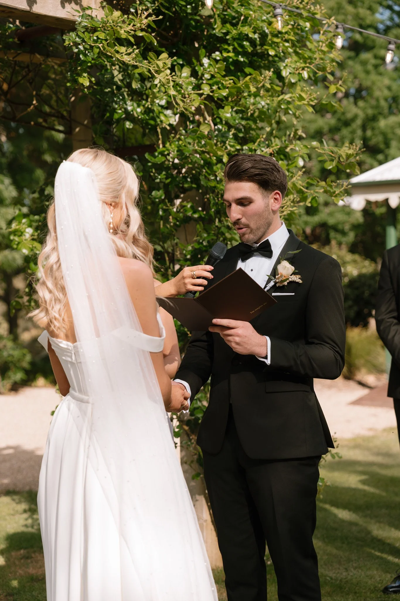 A bride and groom exchange vows outdoors during their wedding ceremony. The bride has long blonde hair and wears a white wedding dress with a veil. The groom, dressed in a black tuxedo with a bow tie, holds a book and is speaking as they shake hands.
