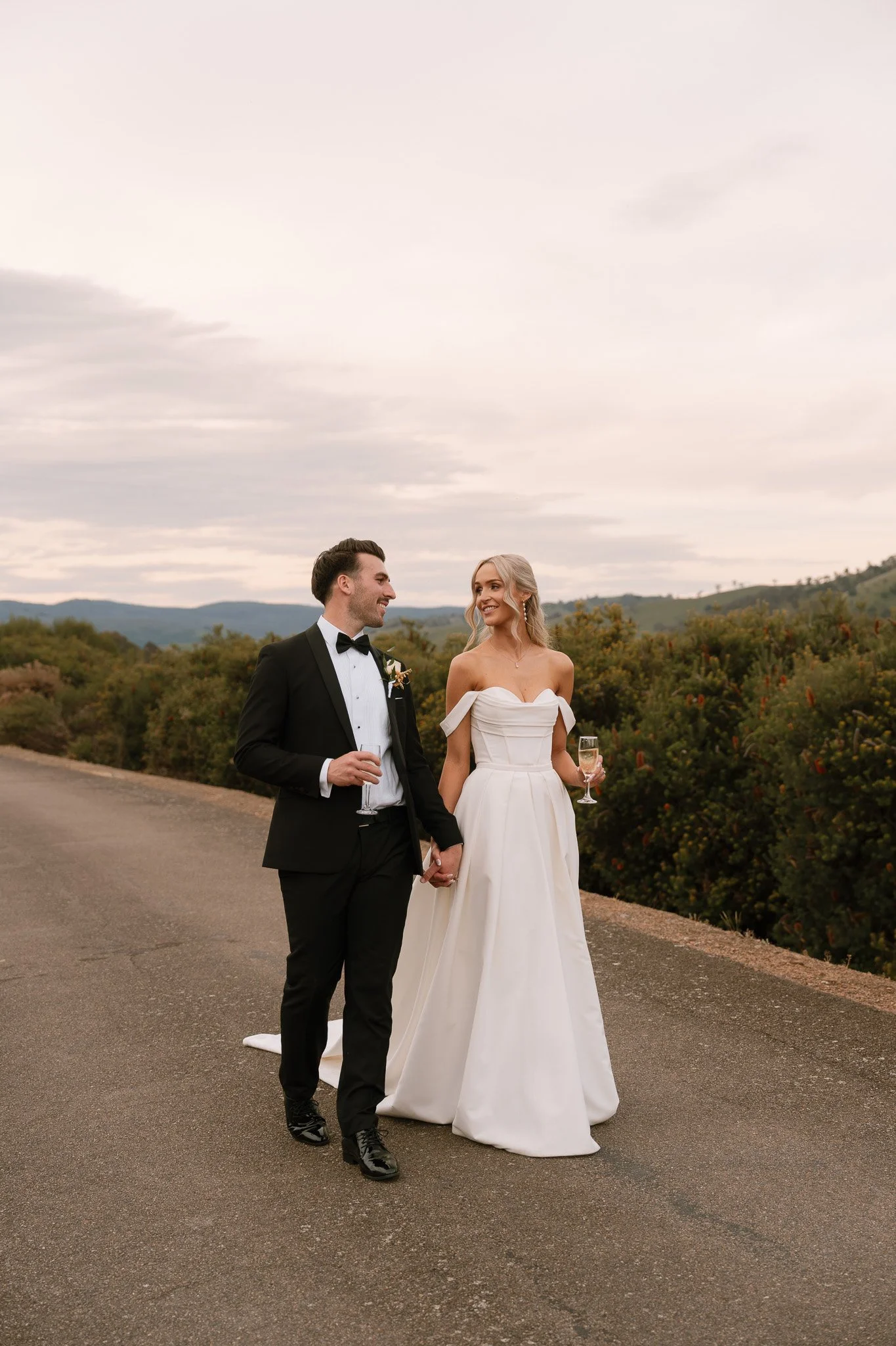 A newlywed couple is walking hand-in-hand on a paved road in a scenic outdoor setting, with the groom in a black tuxedo and the bride in a white wedding gown, holding glasses of champagne and smiling at each other.