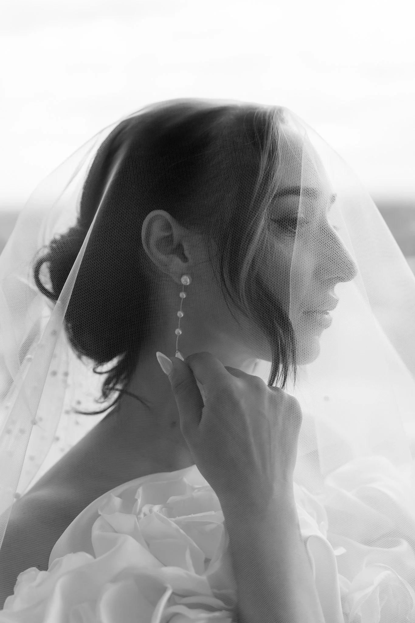 A black and white portrait of a woman wearing a veil and earrings, with her hand resting near her chin.