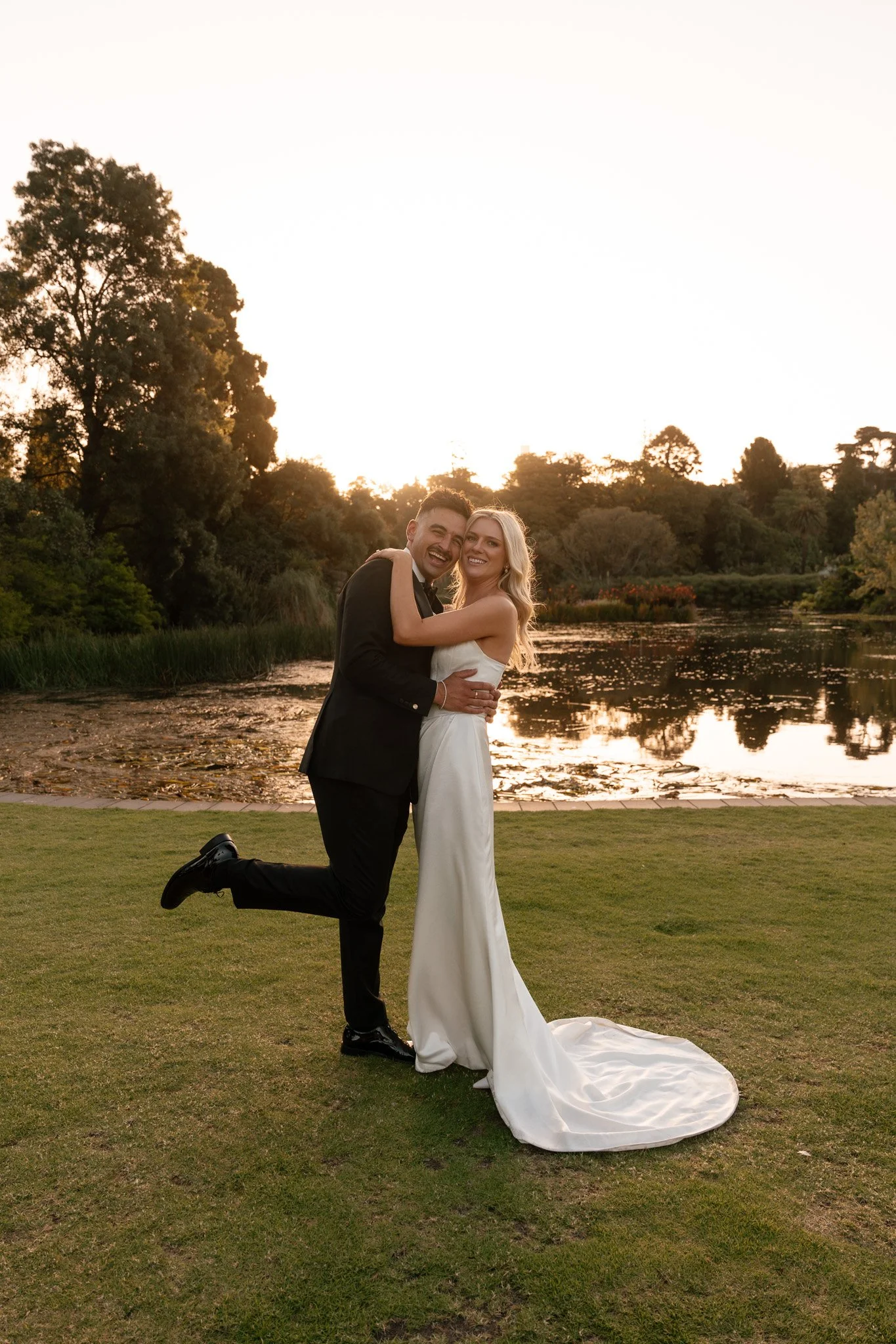 A happy couple in wedding attire embracing near a pond at sunset, with trees and a clear sky in the background.