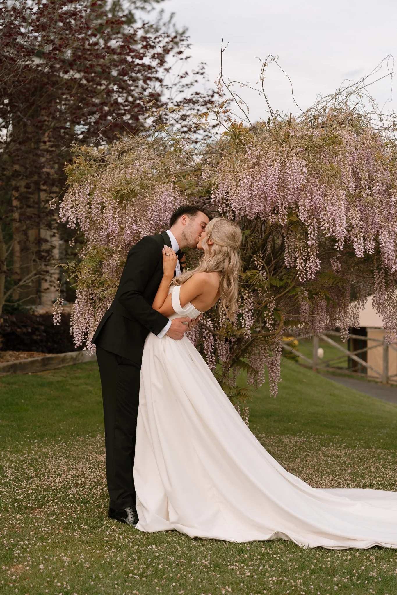 A bride and groom kissing in front of a pink flowering tree during their wedding photoshoot.