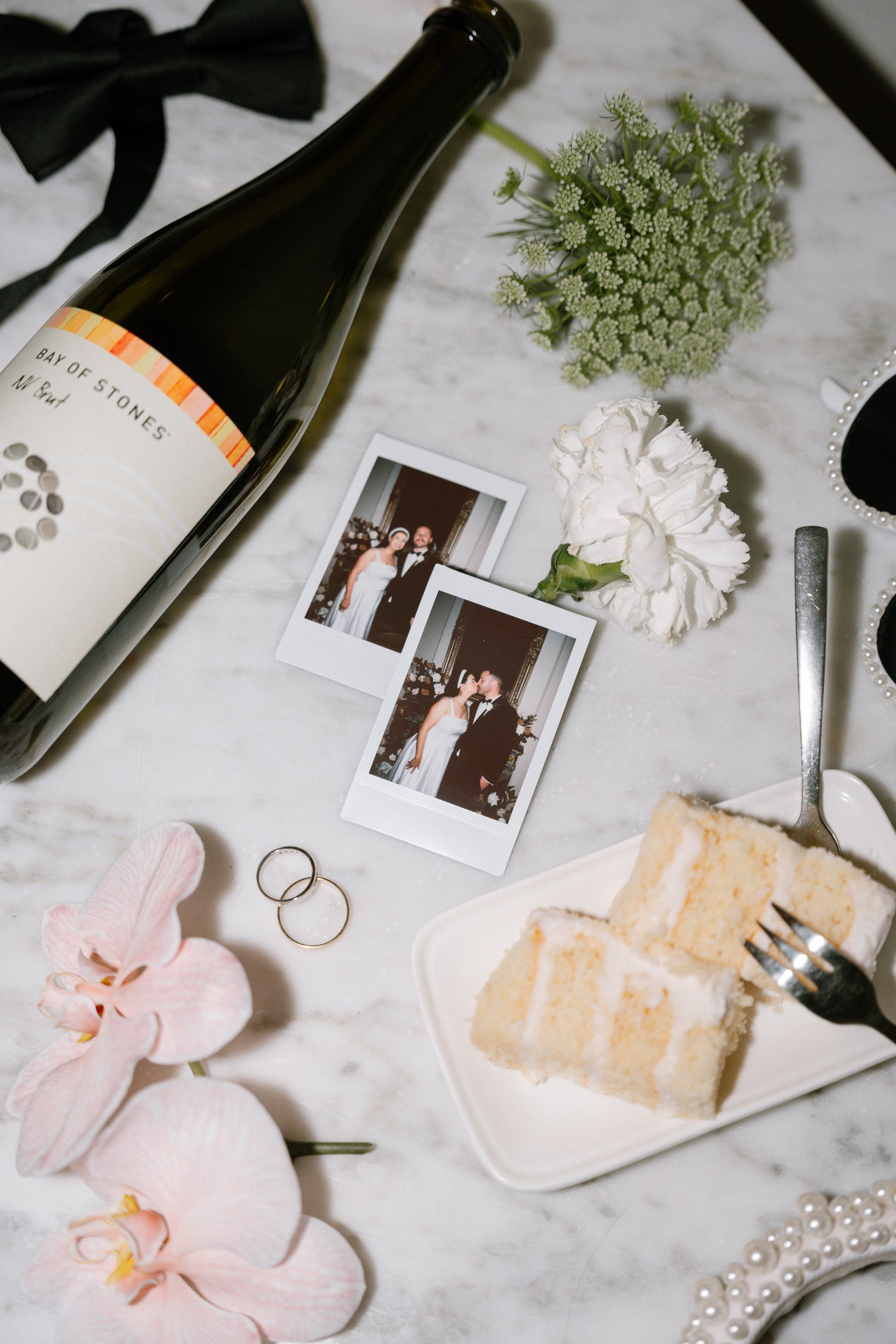 Wedding-themed flat lay with champagne bottle, cake slices, flowers, wedding rings, polaroid photos, and a bow tie on marble background.