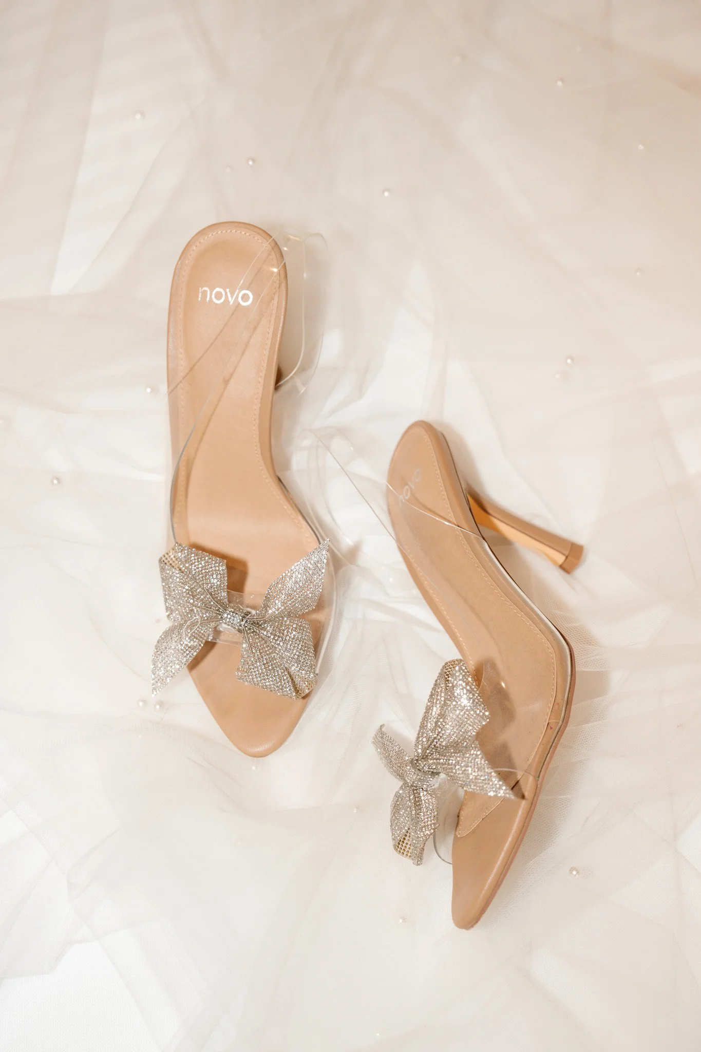 Beige high-heeled shoes with large rhinestone bows on the front, placed on a sheer white fabric background.
