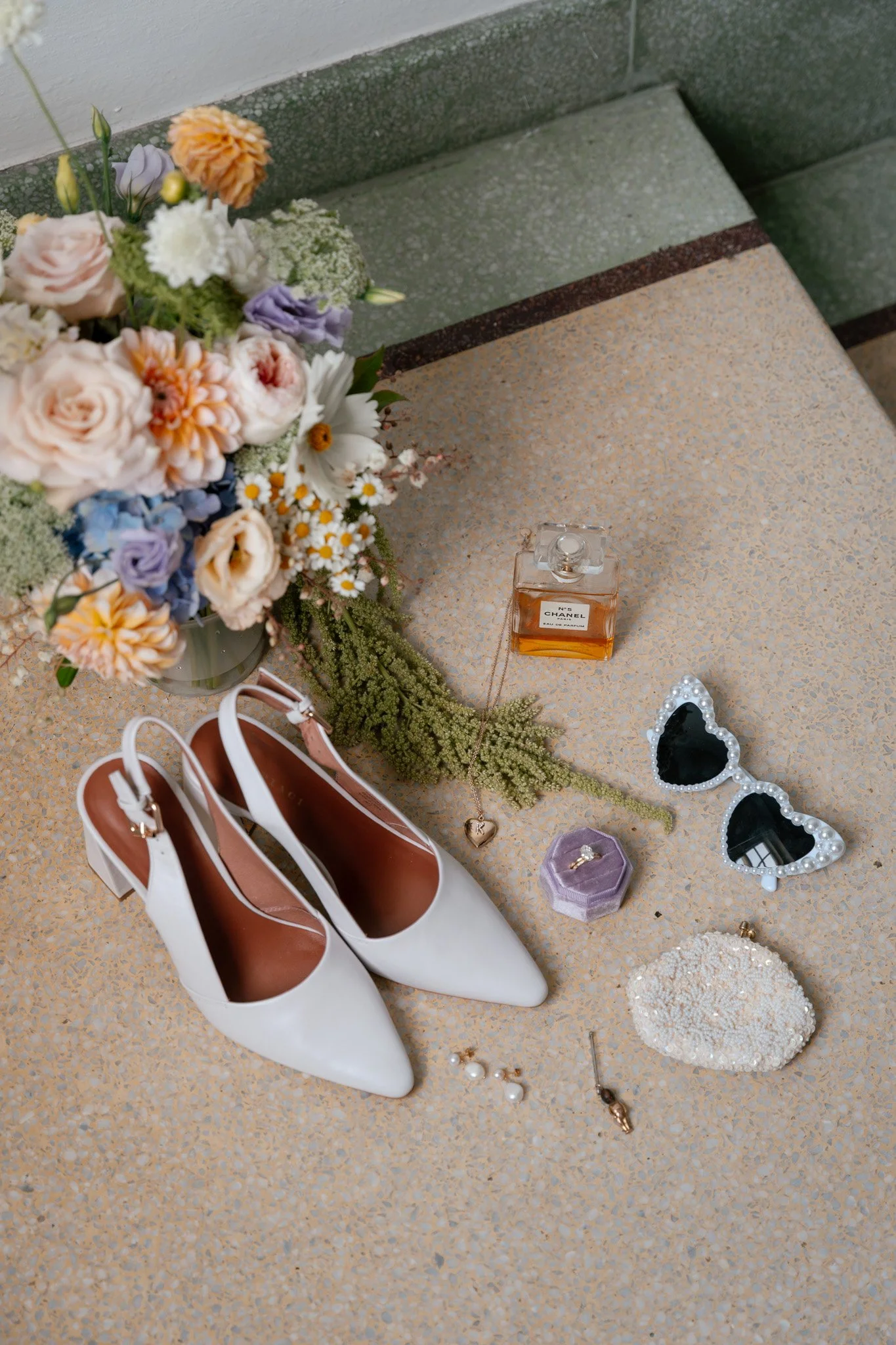 A collection of wedding accessories including a bouquet of flowers, white high-heeled shoes, a bottle of Chanel perfume, a pair of heart-shaped sunglasses with pearl embellishments, a jewelry box with a ring, a beaded clutch, earrings, a necklace wit