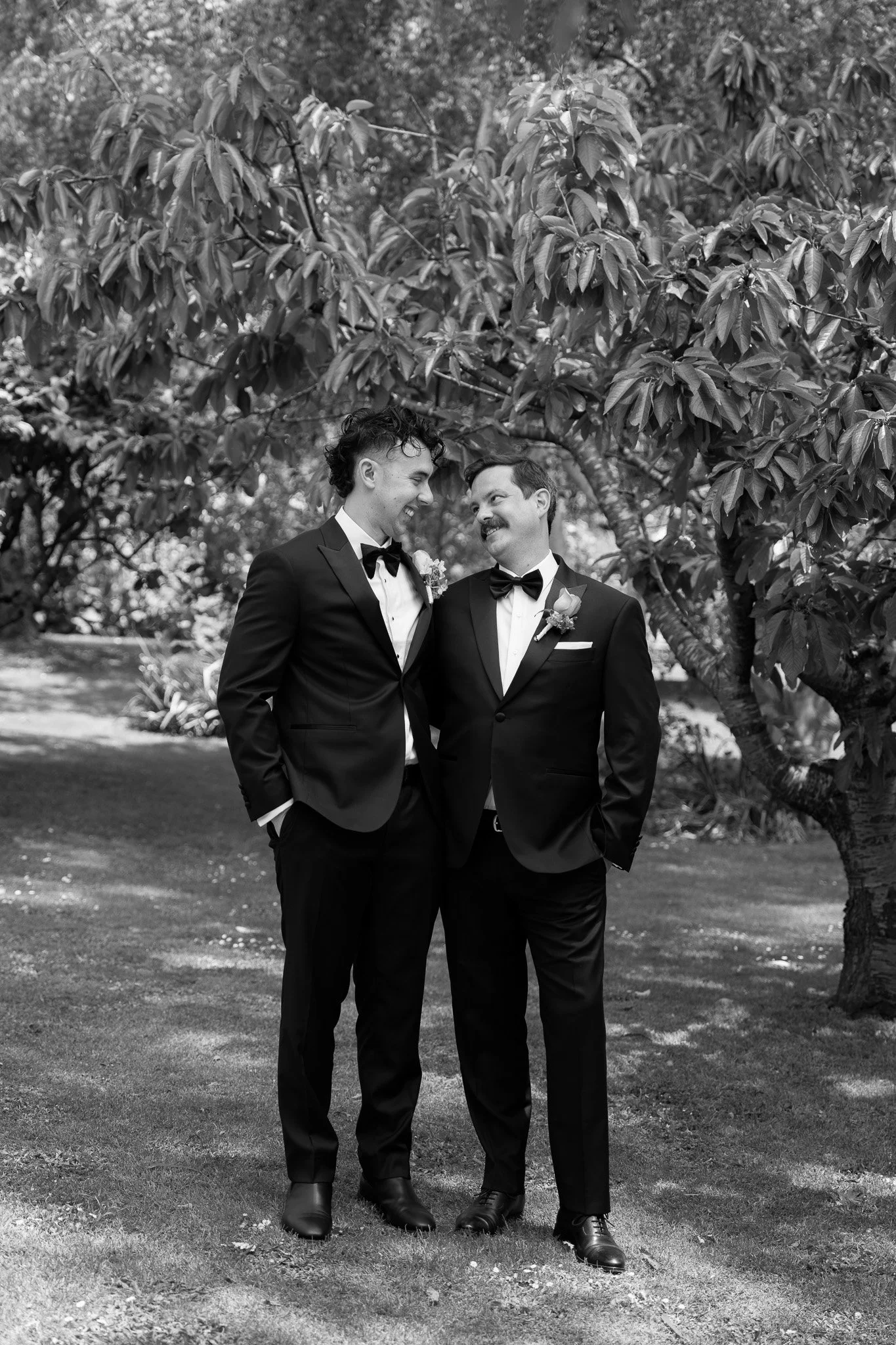 Black and white photo of two men in tuxedos standing outdoors under a large leafy tree, smiling and looking at each other.
