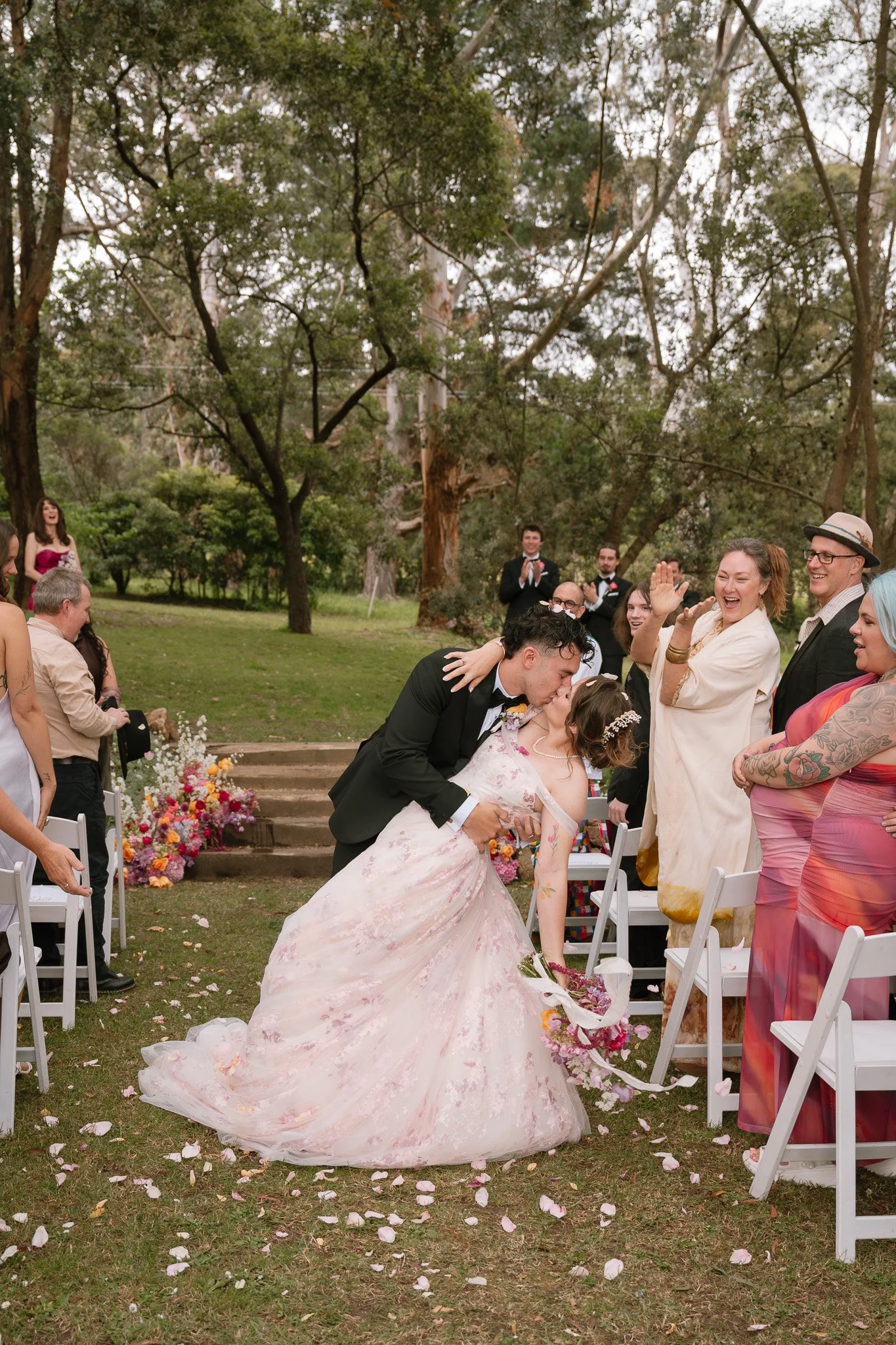 A wedding ceremony outdoors with the bride and groom sharing a kiss. The bride is in a wedding gown, and the groom in a tuxedo. Guests are clapping and celebrating, surrounded by trees and flower decorations.