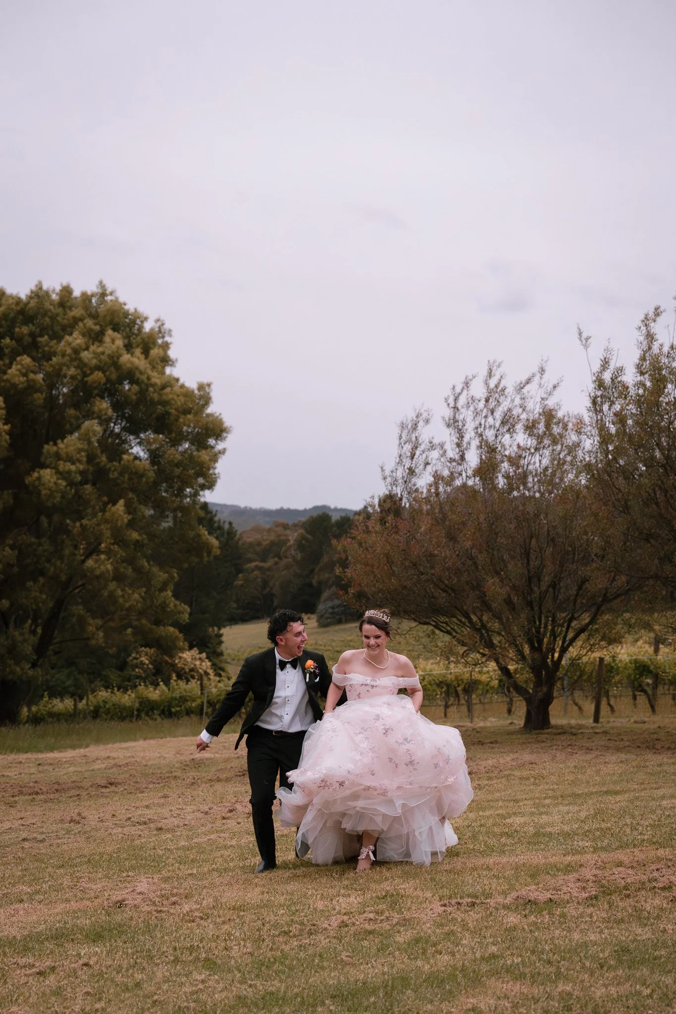 A bride and groom running together outdoors in a field, smiling and laughing. The bride is wearing a pink floral wedding dress and a tiara, while the groom is in a black tuxedo with a bow tie.