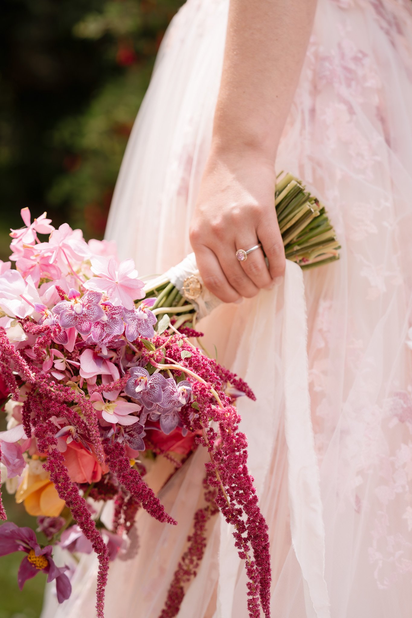 Close-up of a woman in a light pink dress holding a bouquet of pink and purple flowers, with an engagement ring on her finger.