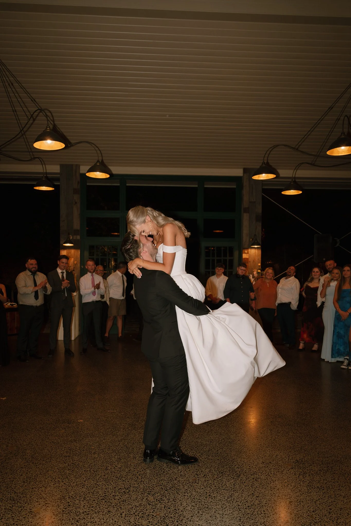 A bride and groom dance at their wedding reception, with guests watching and clapping in the background inside a decorated venue.