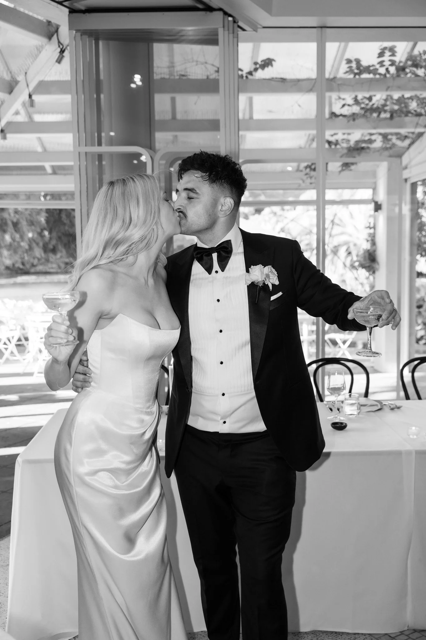 A couple in formal wedding attire sharing a kiss at a reception, holding drinks, with a decorated table in the background.