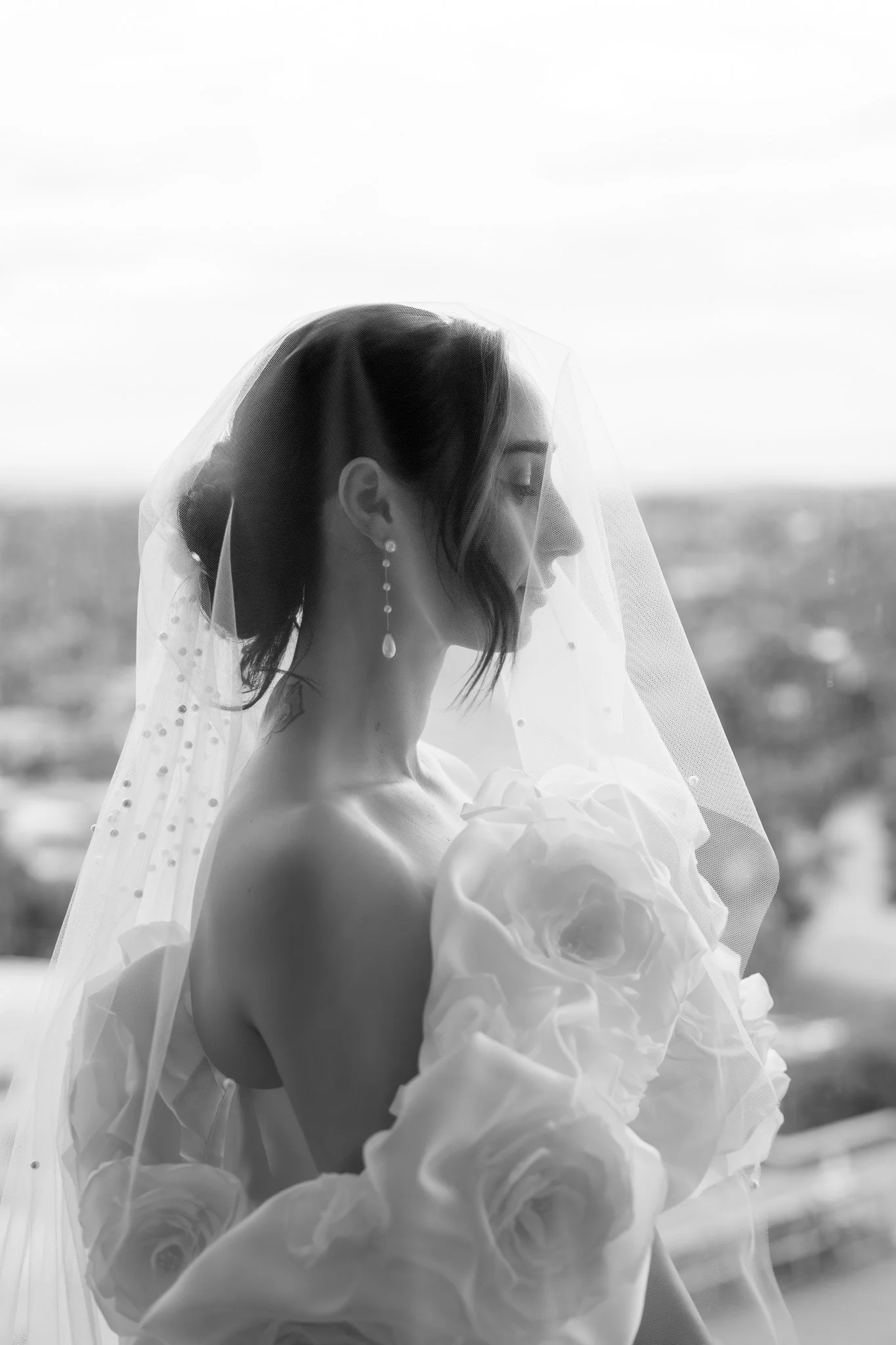 Black and white photo of a bride with eyes closed, wearing a veil and earrings, holding a bouquet of large roses.