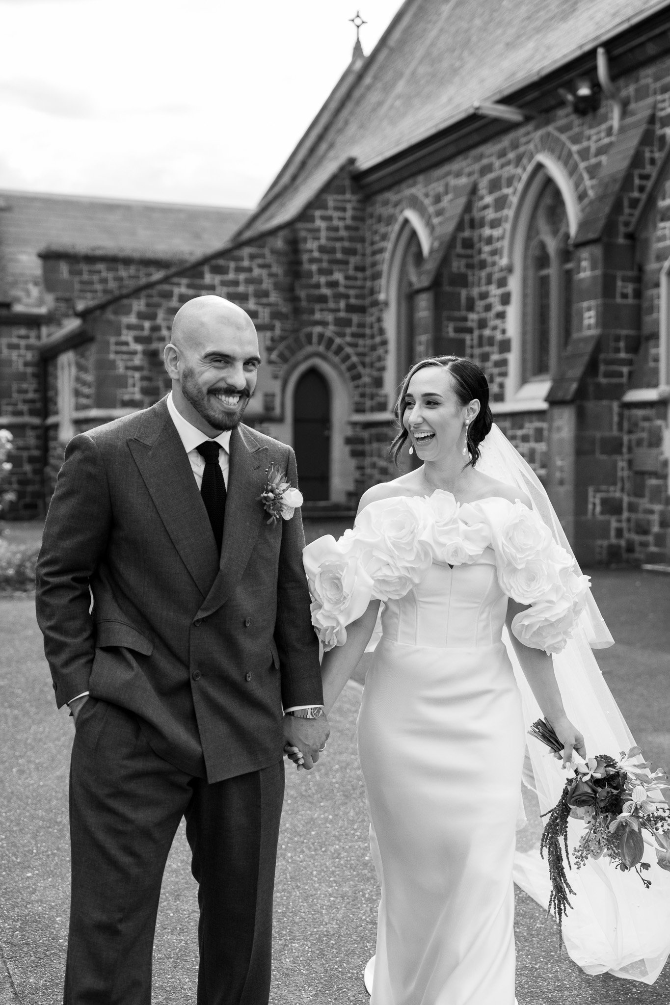 Black and white photo of a smiling bride and groom holding hands outside a church.