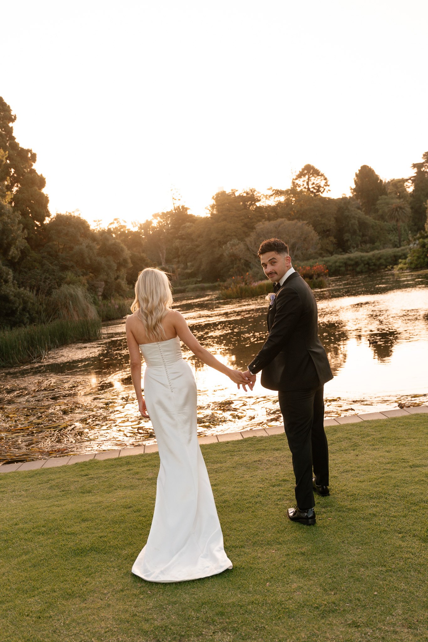 A newlywed couple holding hands by a lake during sunset. The bride wears a strapless white gown with front buttons, and the groom wears a black tuxedo. The background features trees and a calm lake reflecting the sky.