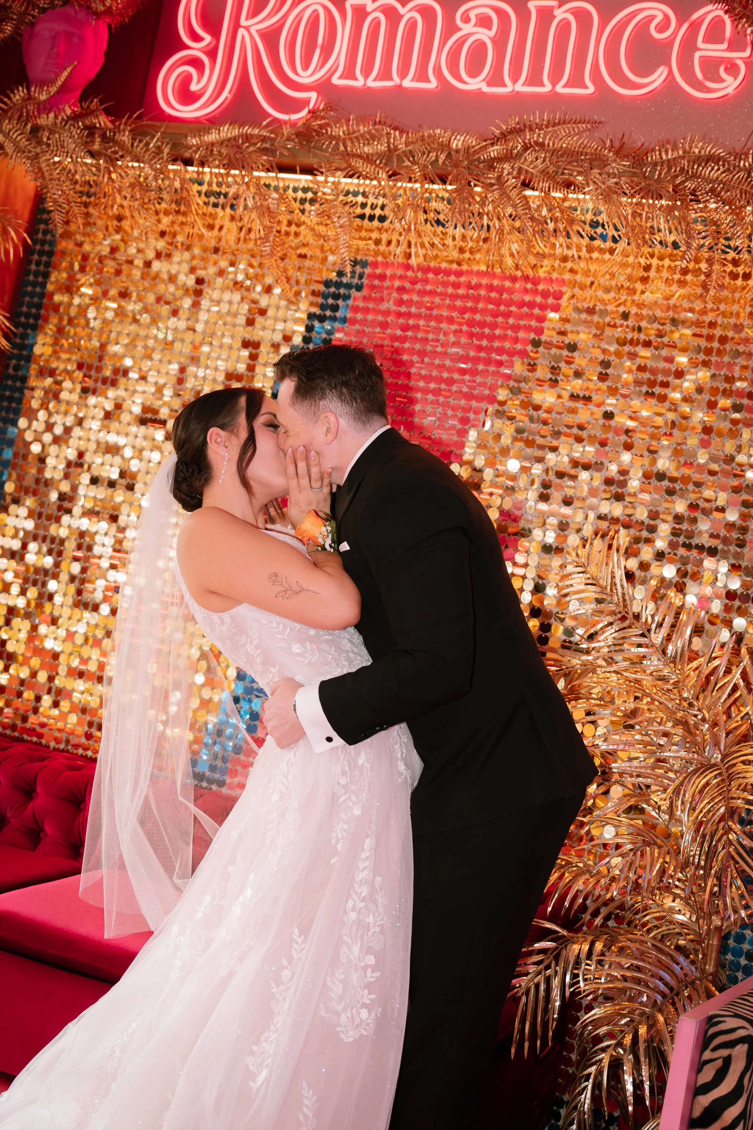 A bride and groom sharing a kiss during their wedding reception, with a glamorous background sign reading 'Romance' and decorated with golden leaves and shiny, circular decorations.