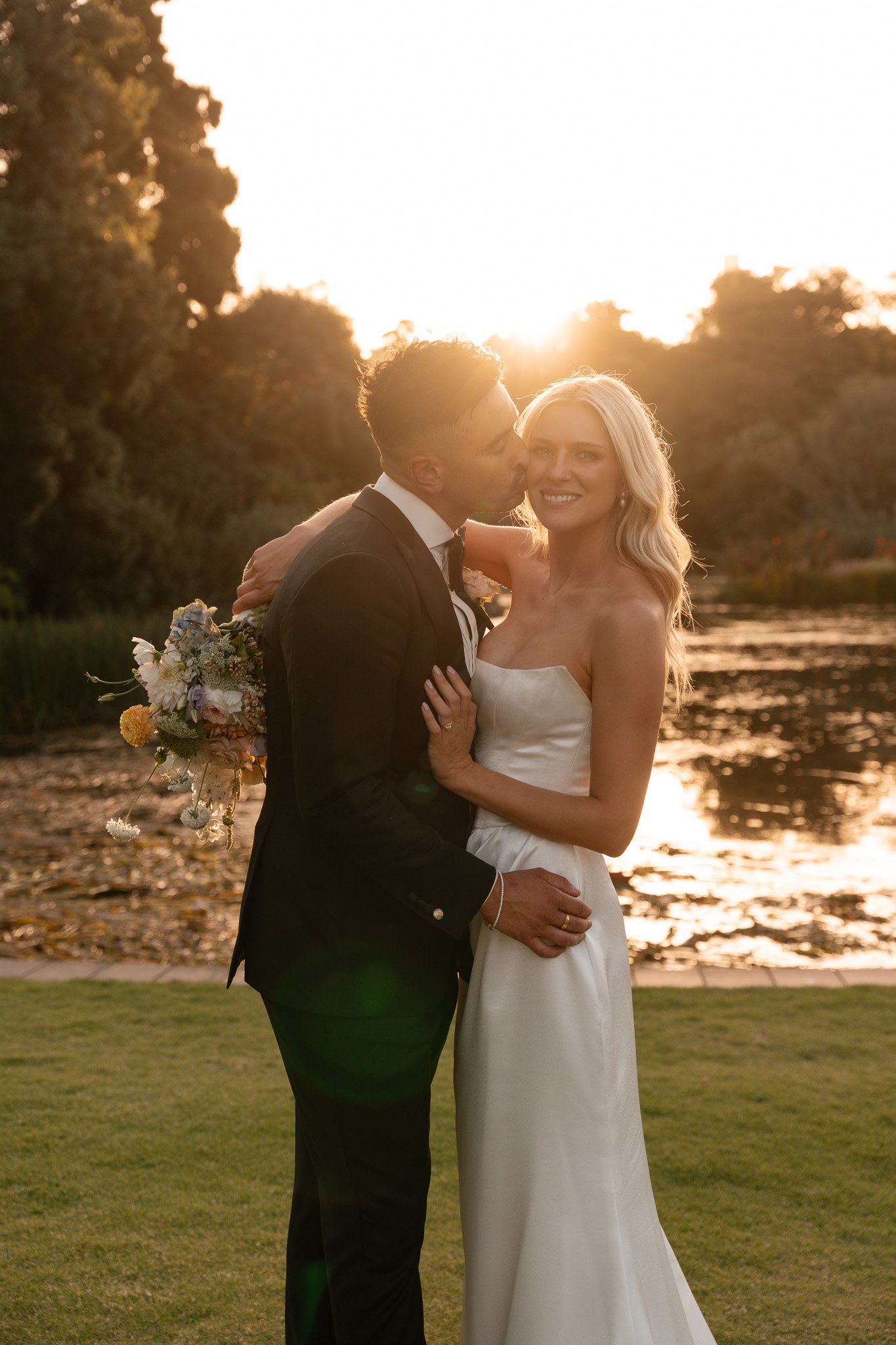 A newlywed couple standing outdoors near a river at sunset, with the bride in a white wedding dress and the groom in a black suit. The groom is kissing the bride on her cheek, and they are smiling. The bride is holding a bouquet of flowers.