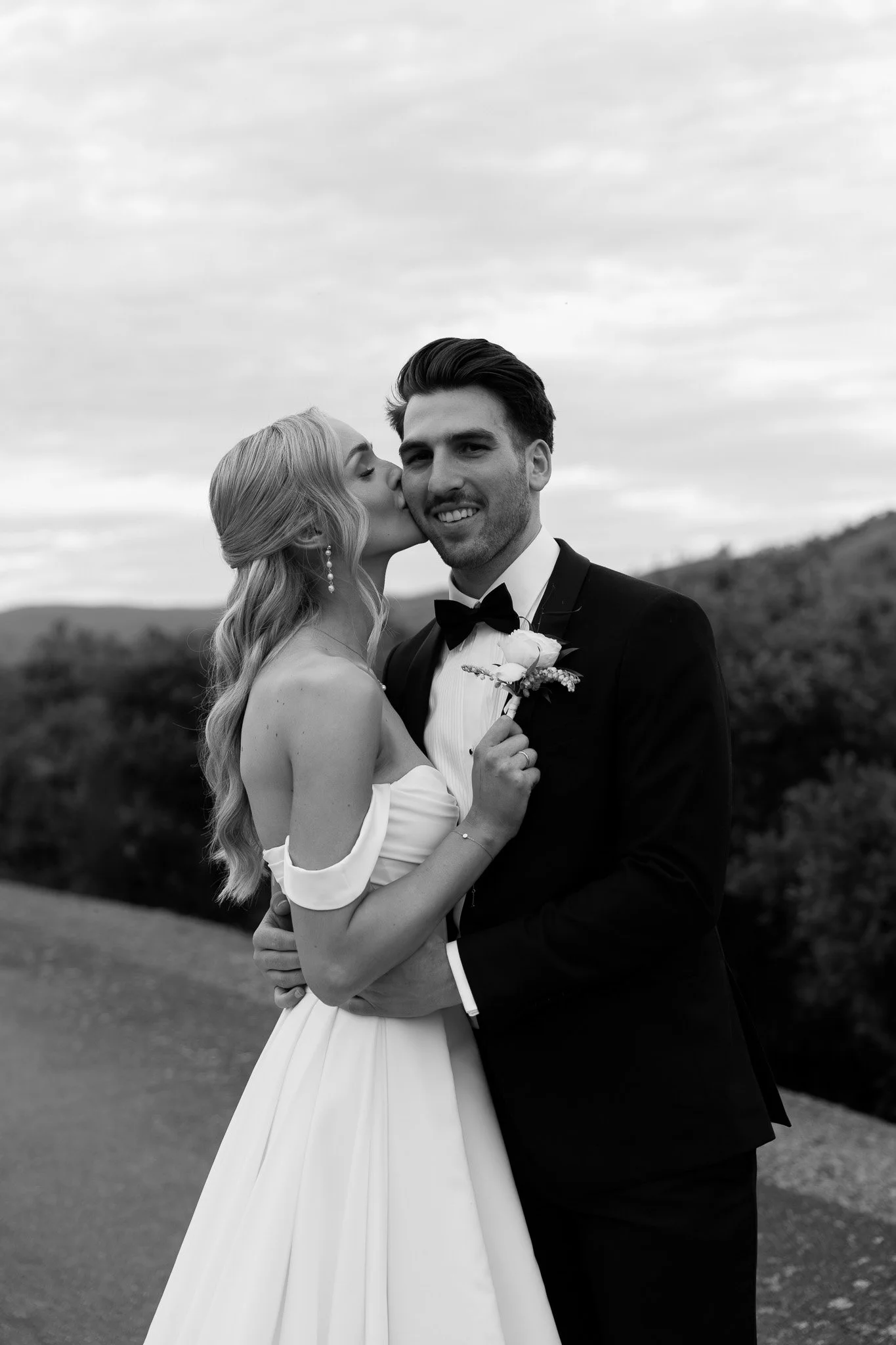 Black and white photo of a bride and groom on their wedding day, with the bride kissing the groom's cheek, and the groom smiling, outdoors with hills and cloudy sky in the background.
