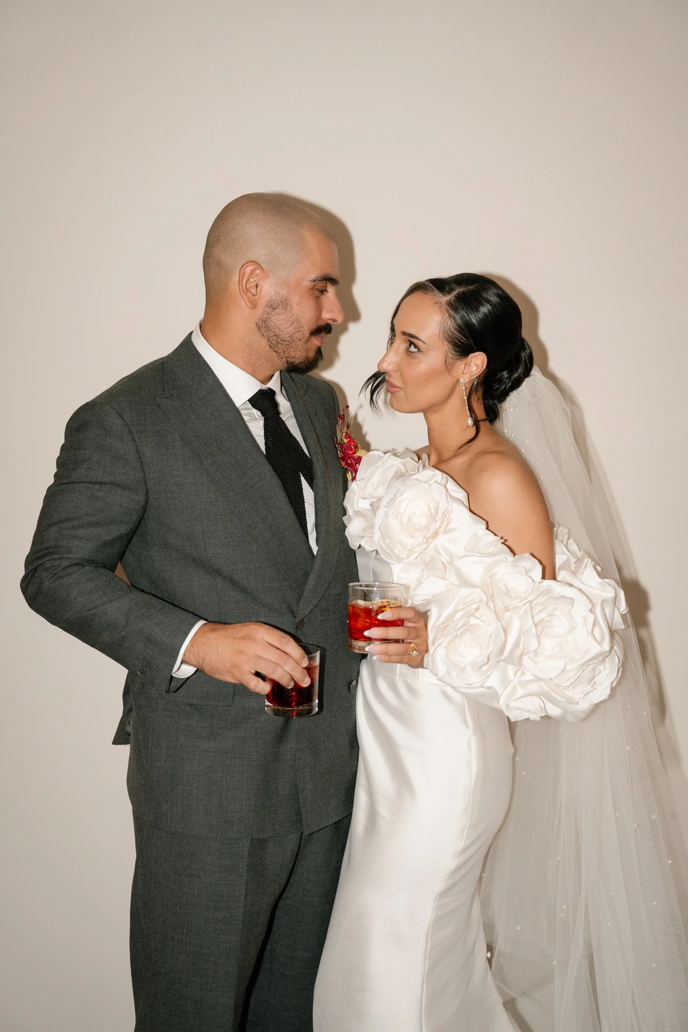 A bride and groom holding glasses of red drink, standing close together, looking into each other's eyes, with a simple white background.