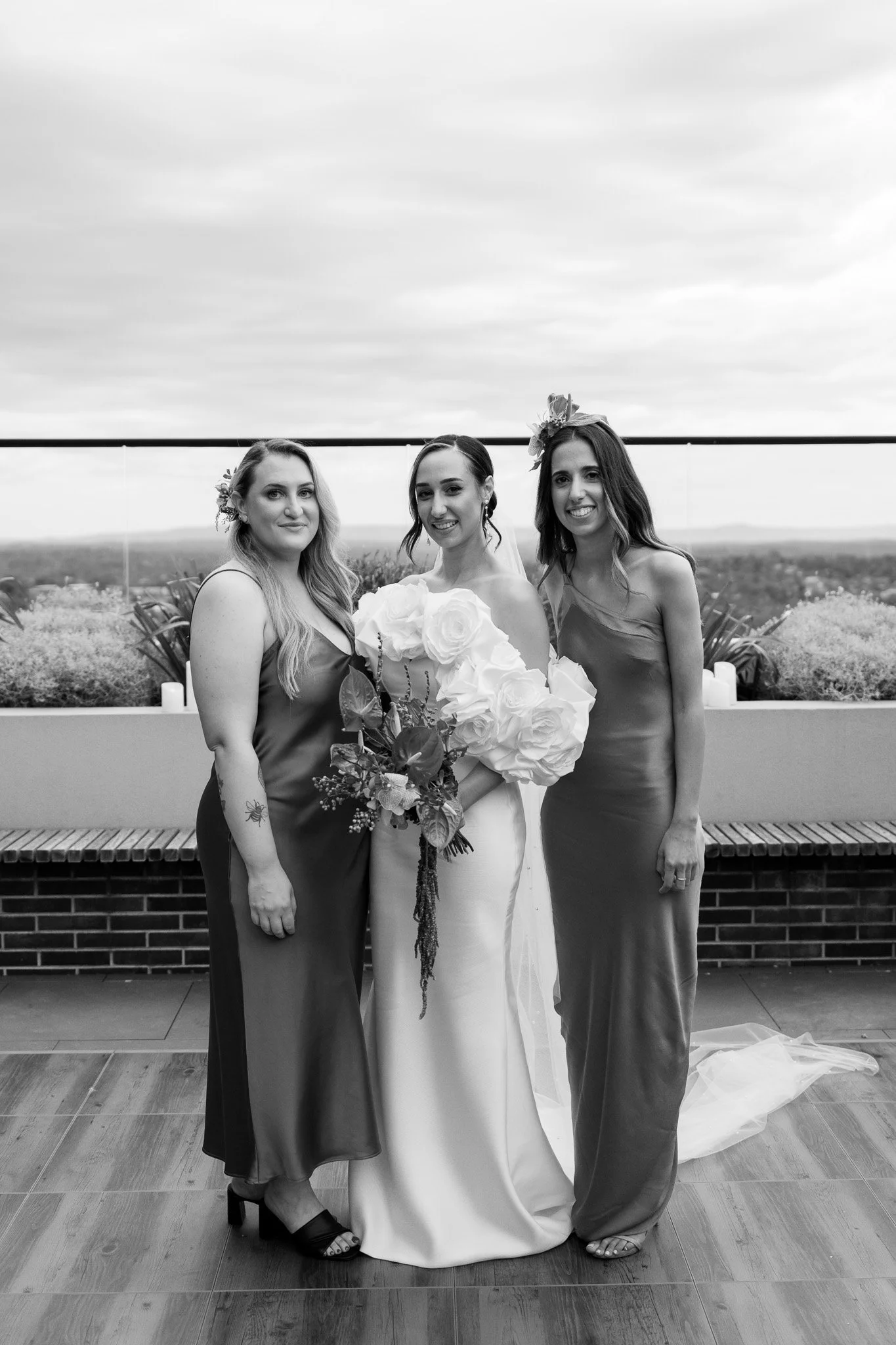 Black and white photo of three women, with one in a white wedding dress holding a bouquet, standing between two women in bridesmaid dresses on a rooftop or outdoor terrace.