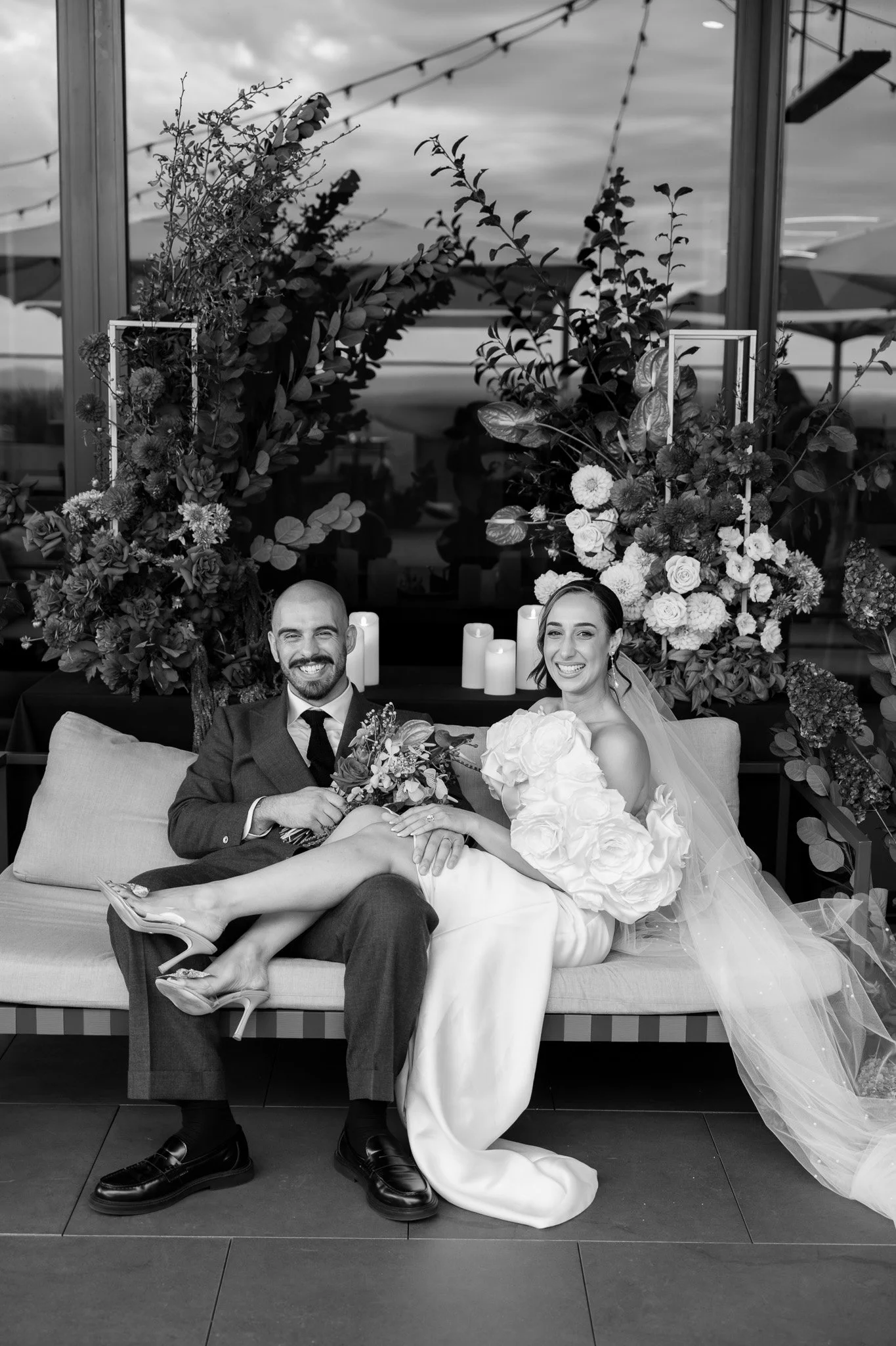 A newlywed couple sitting on a couch, smiling. The man is in a suit, and the woman in a wedding dress holding a bouquet. They are surrounded by floral arrangements and candles.