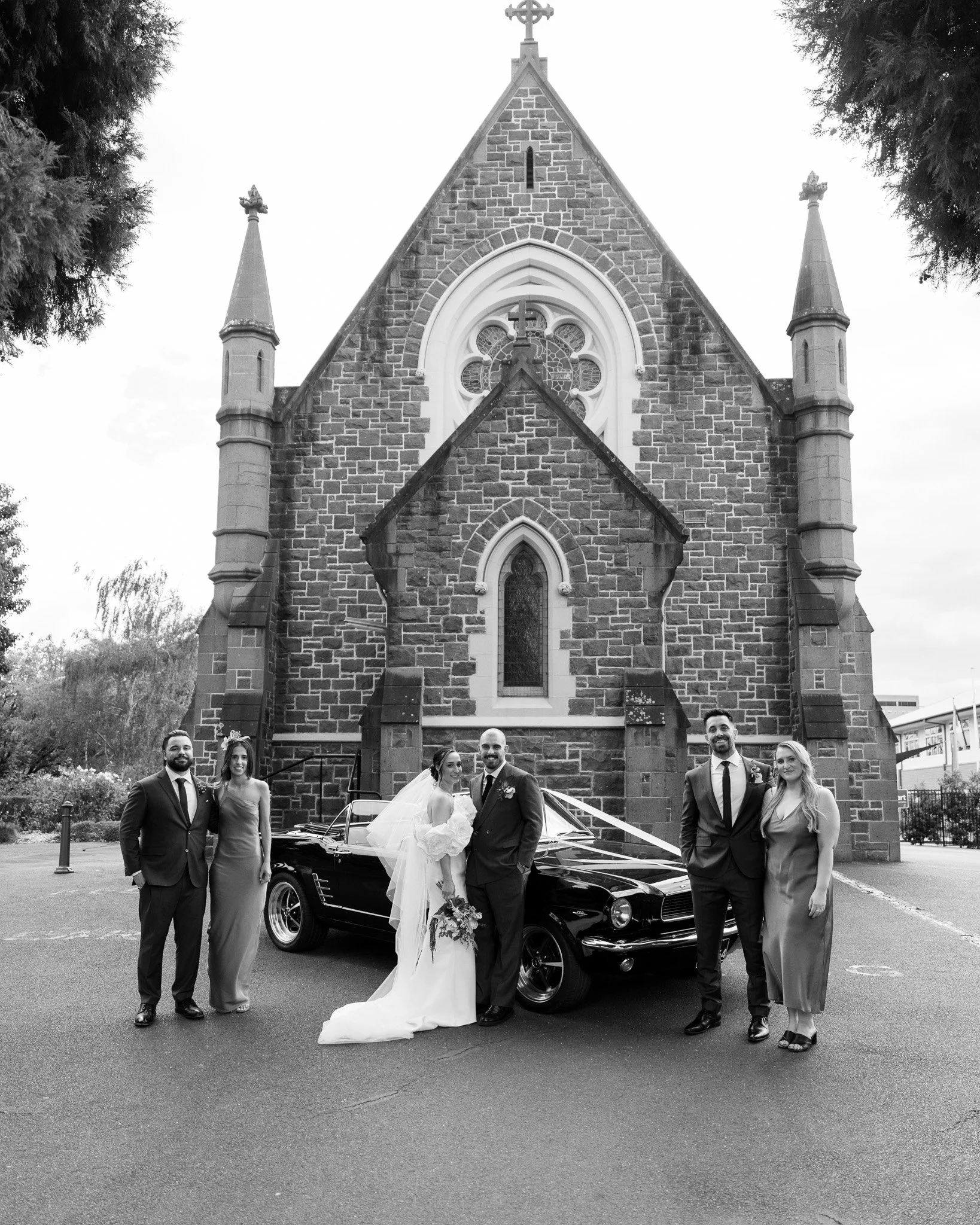 Black and white photo of a wedding party standing in front of a church with a vintage car. The bride and groom are in the center, with the bride holding a bouquet. There are two men and two women, all dressed formally, flanking the couple.