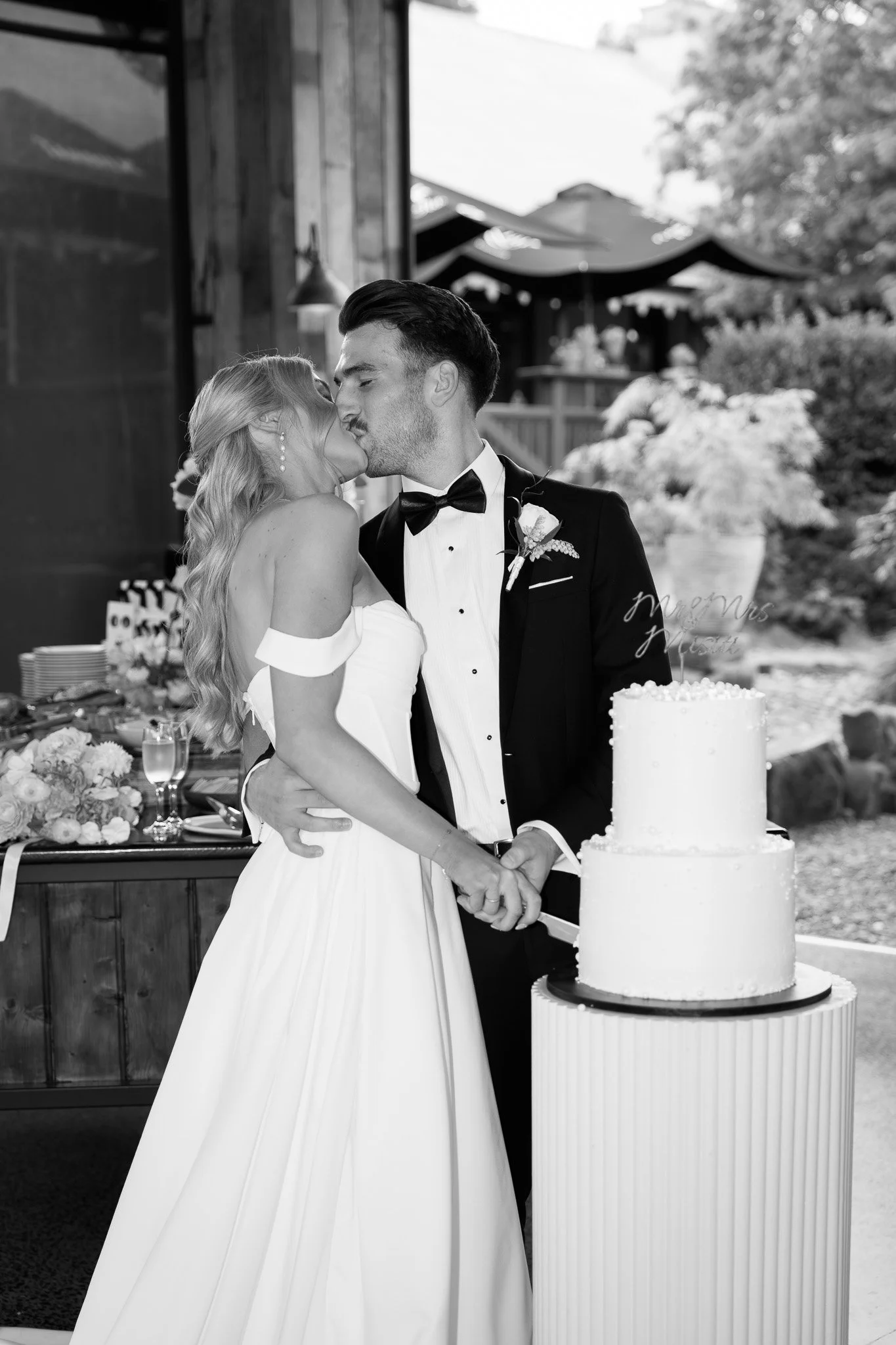 Black and white photo of a newlywed couple kissing while cutting a wedding cake outdoors.