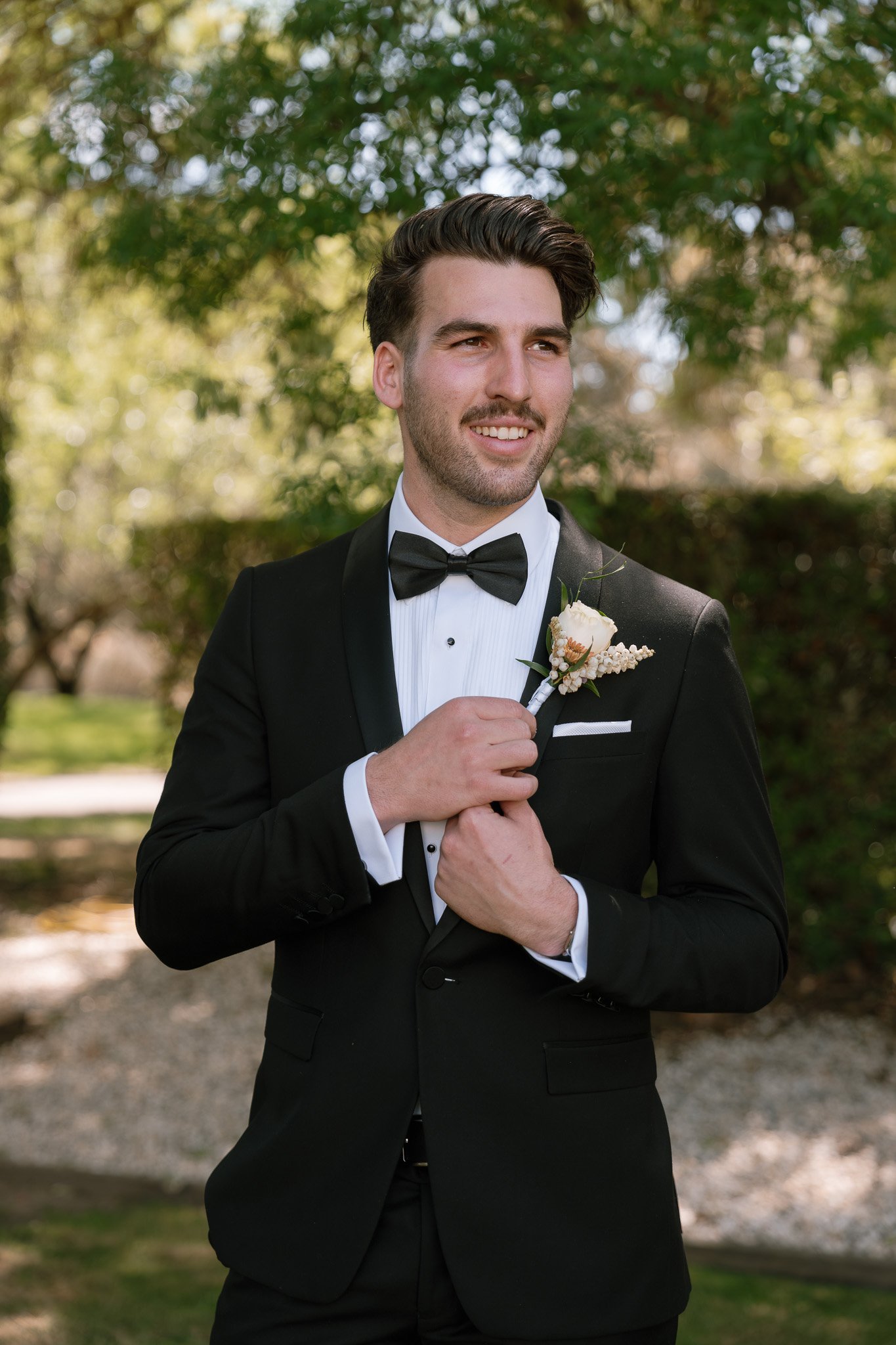 A groom wearing a black tuxedo, white dress shirt, and black bow tie standing outdoors with trees and greenery in the background, holding a boutonniere.