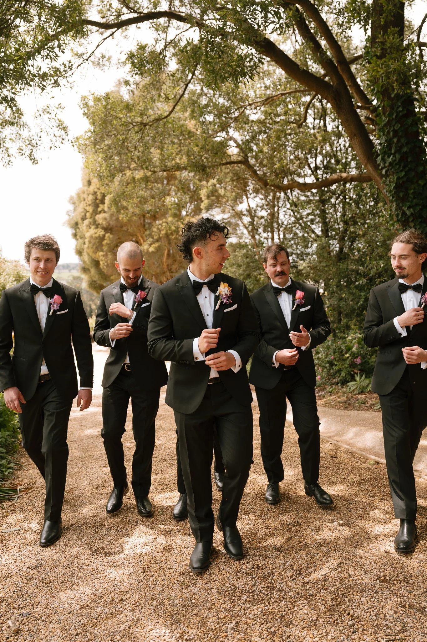 Group of five men in black tuxedos walking outdoors on a garden path, celebrating at a wedding.