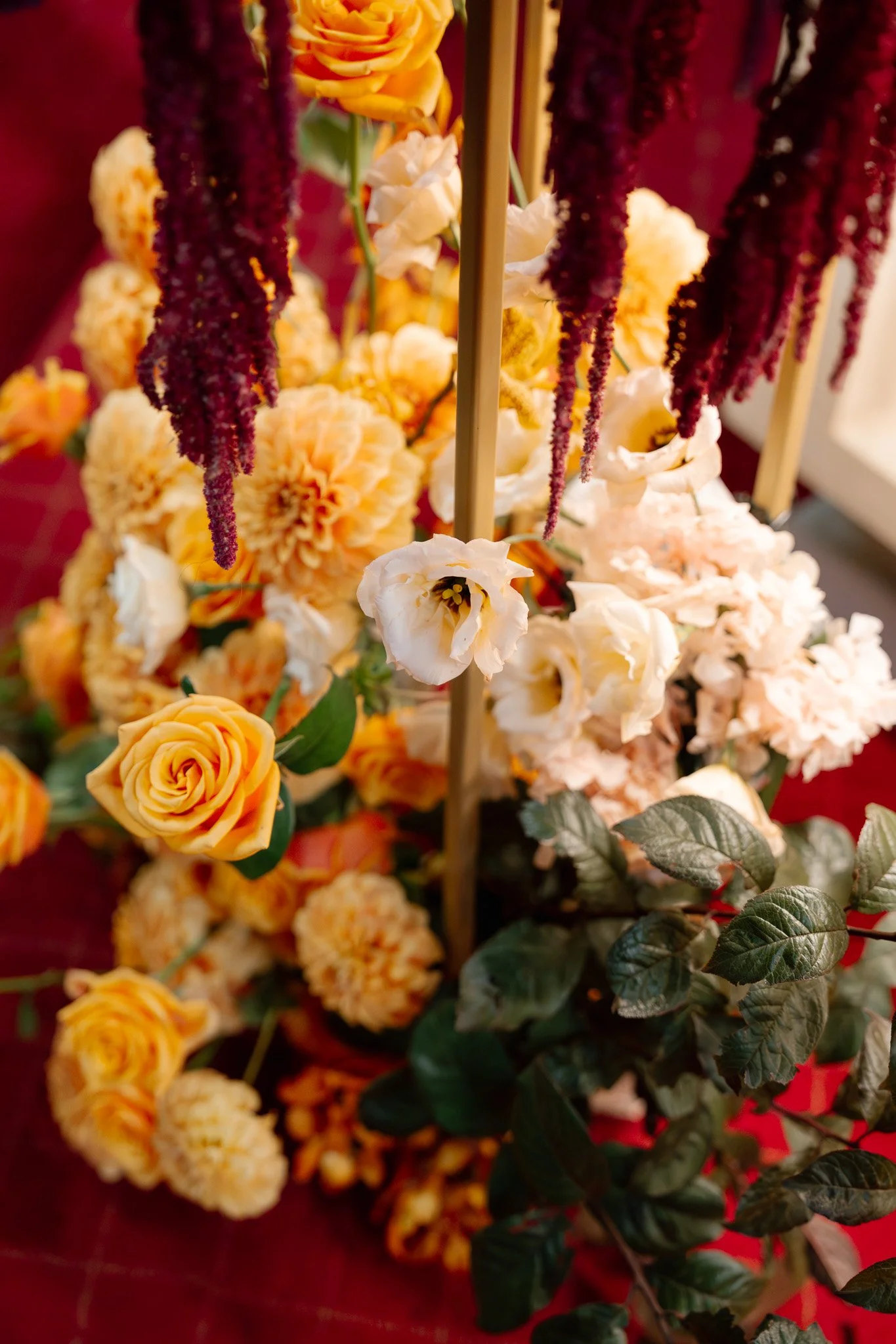A bouquet of yellow roses, cream-colored carnations, white lisianthus, and deep red amaranthus hanging from gold rods, set against a red background.