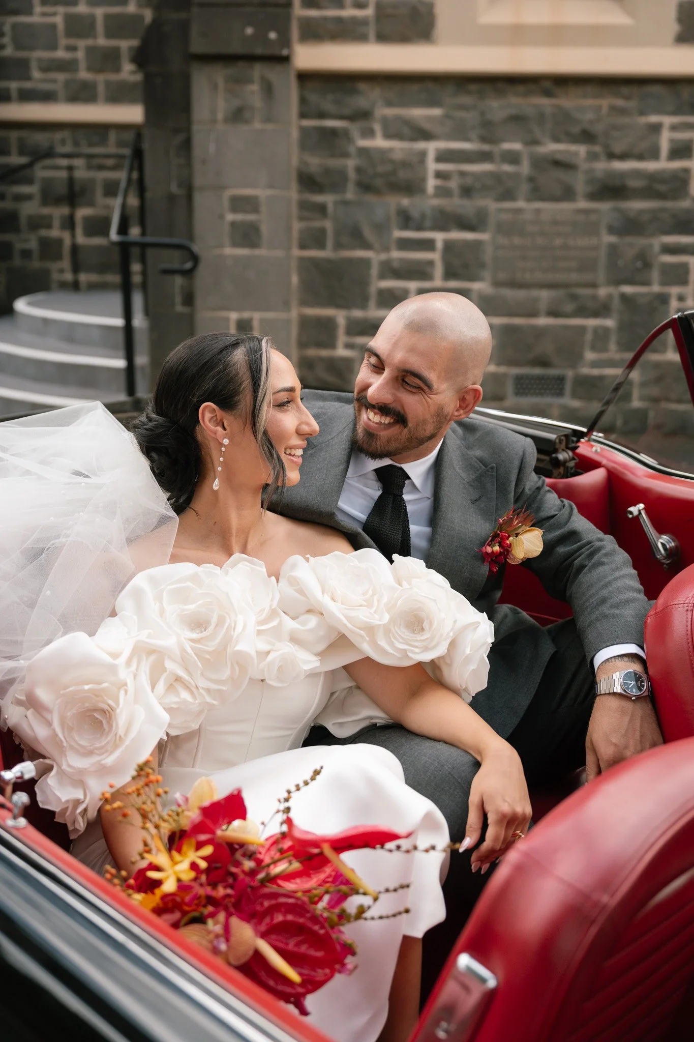 A bride and groom sitting in a vintage red car, smiling and looking at each other, with a stone building background. The bride is wearing a white wedding dress with large floral details on the shoulder, and the groom is dressed in a gray suit with a 