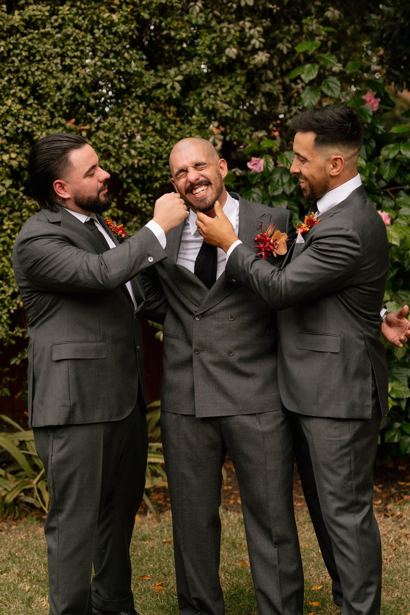 Three men in suits smiling and joking with each other in a garden with greenery and pink flowers in the background.