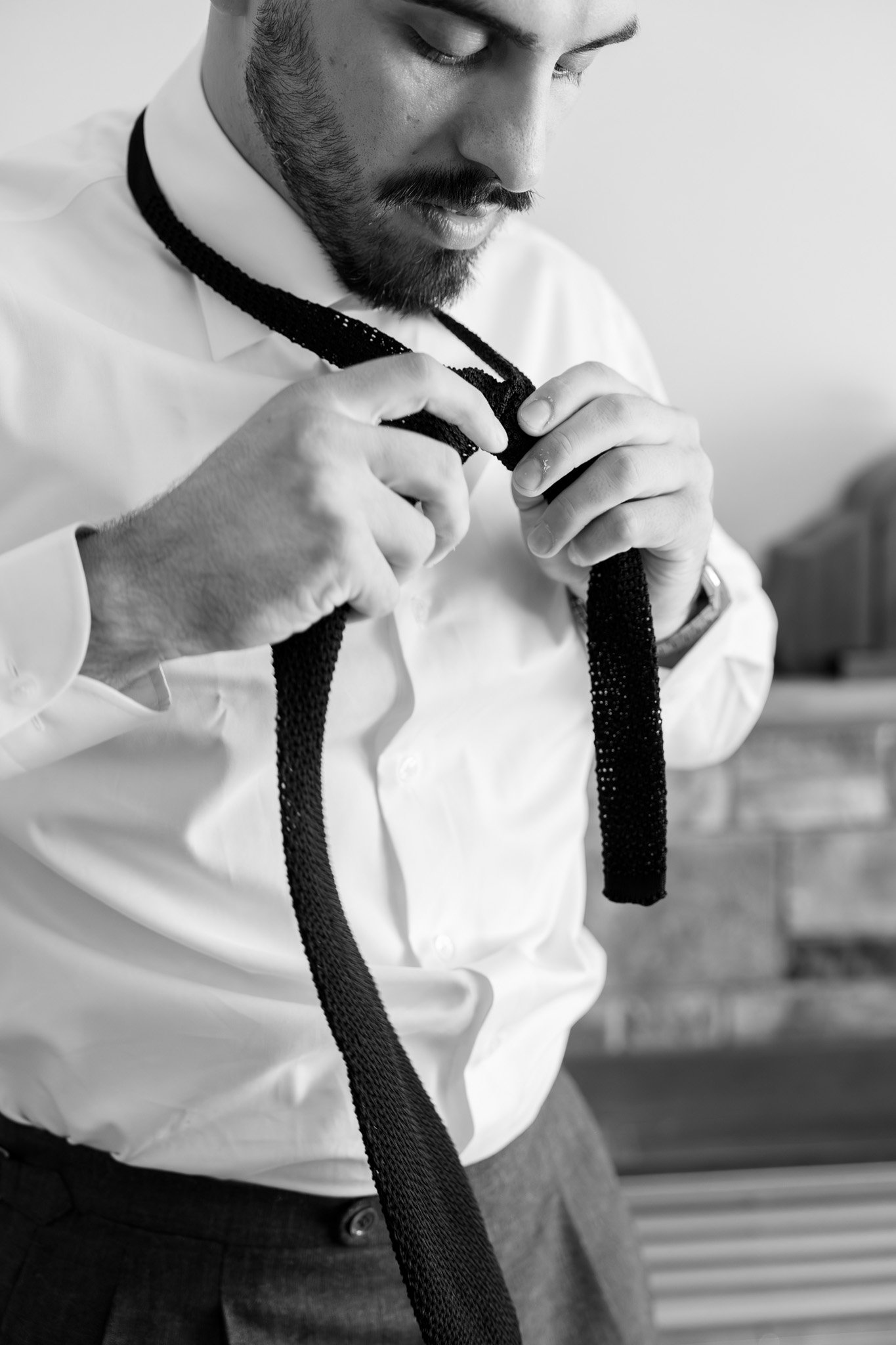 A man in a white shirt is tying a black tie in a room with what appears to be a couch and a table in the background.