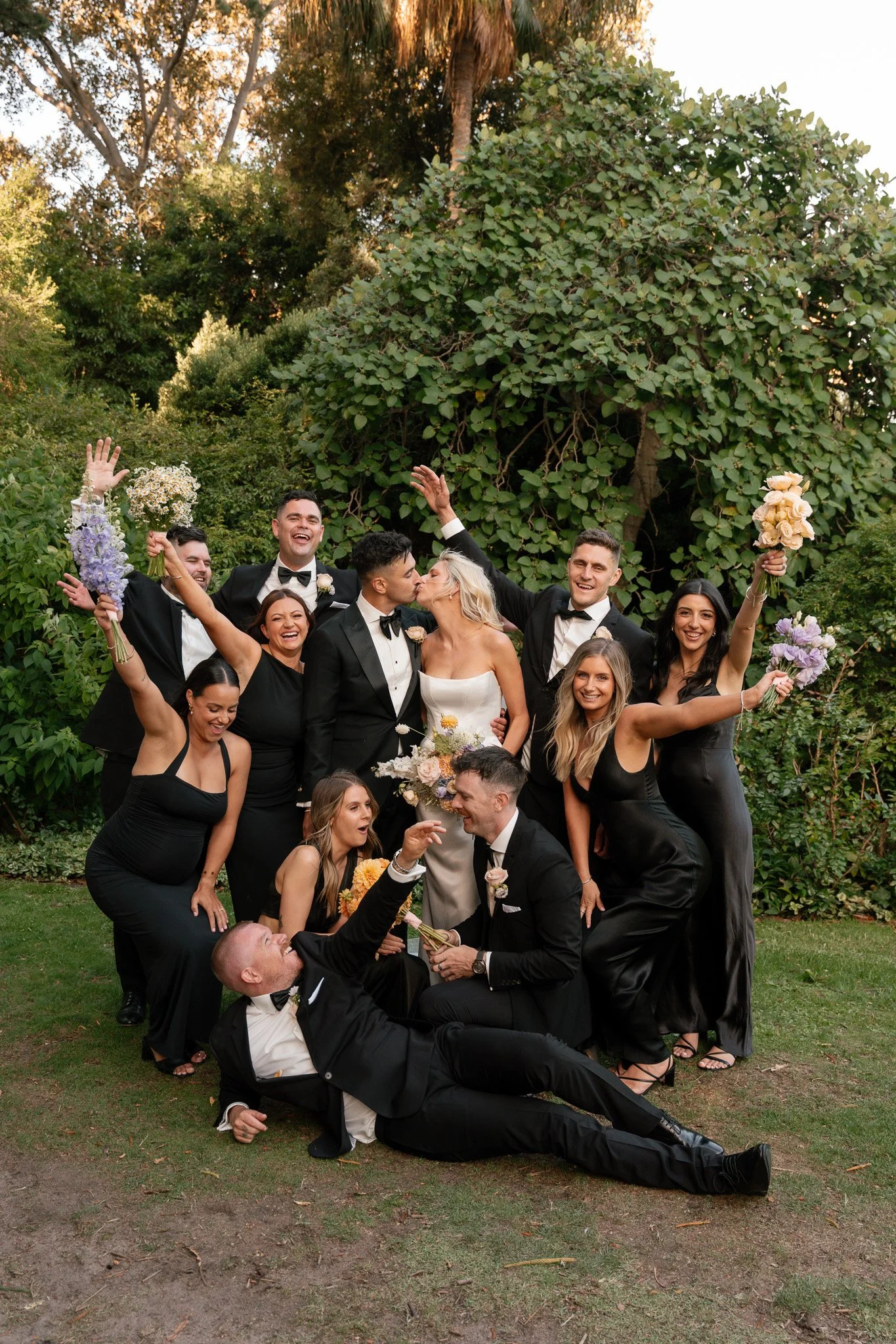 A wedding party celebrating outdoors with the bride and groom in the center, surrounded by friends and family, all dressed in formal black attire, holding bouquets, and smiling happily.