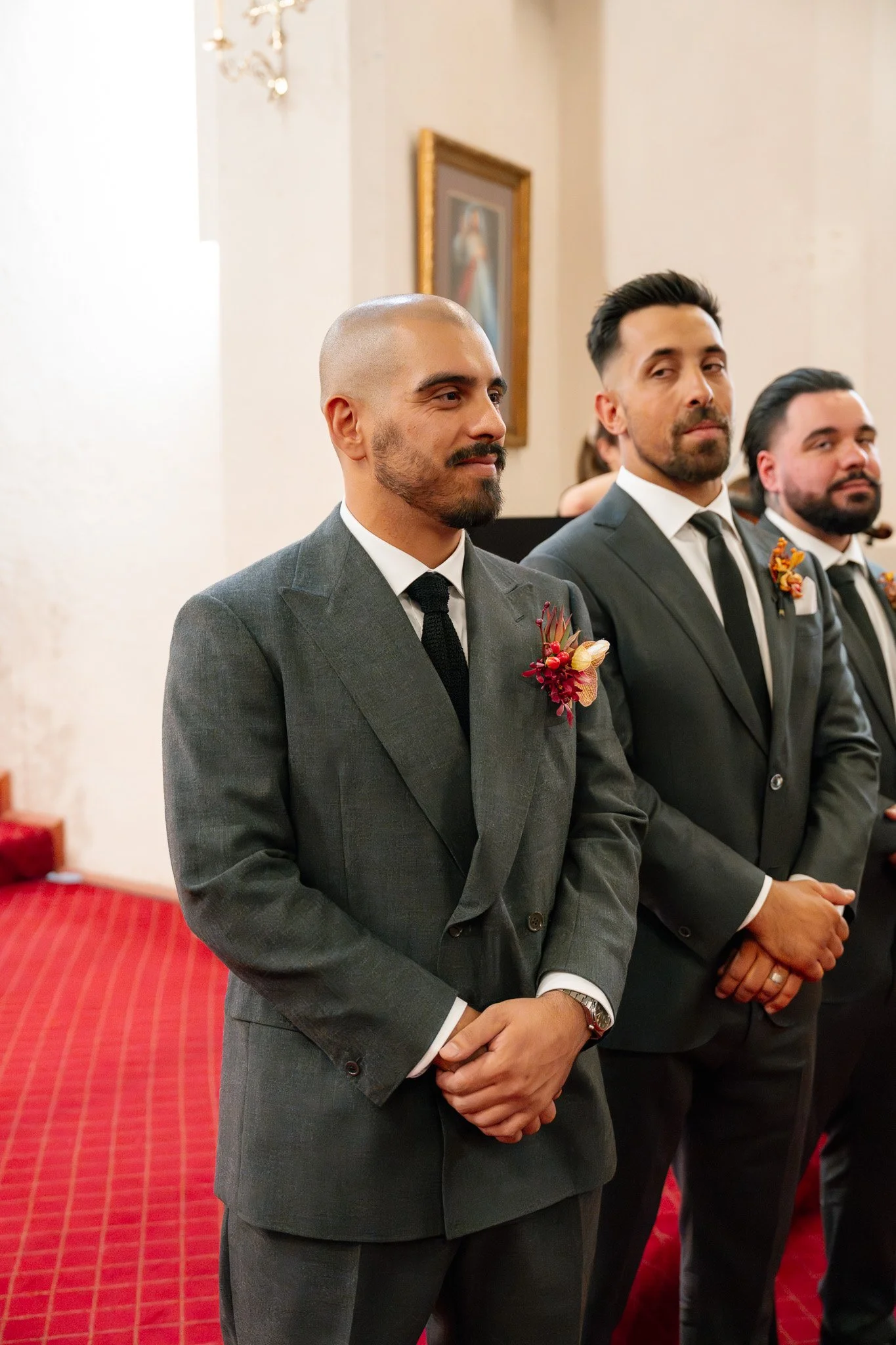 Three men dressed in suits standing during a formal event, likely a wedding, with one in front and the others behind him. All are wearing boutonnières.