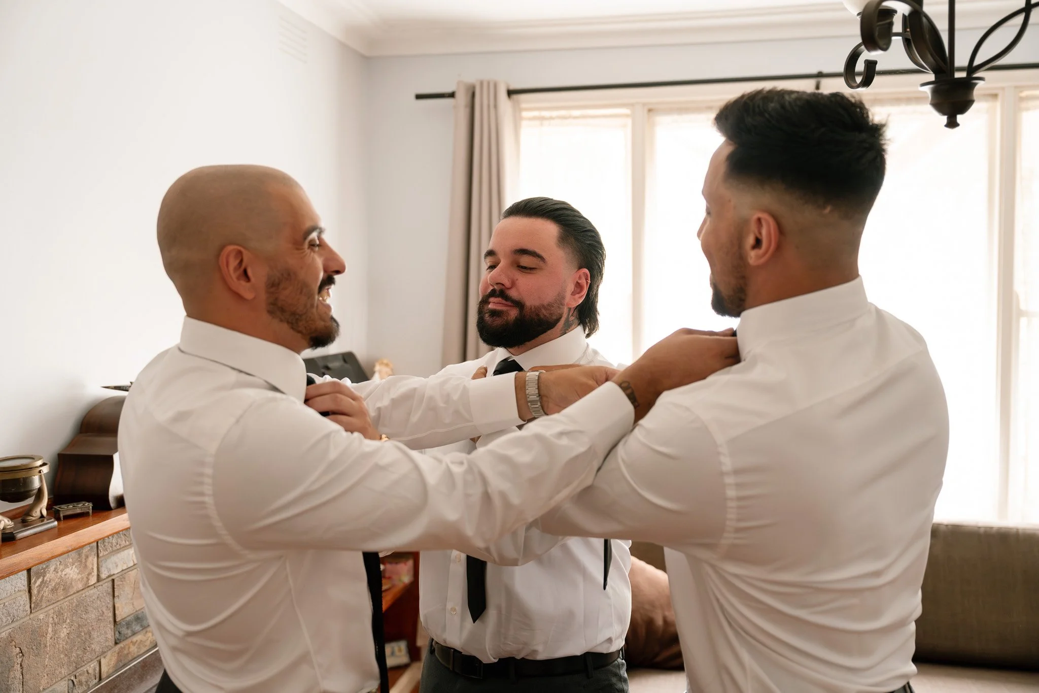 Three men are getting dressed up in white shirts and black ties, assisting each other with their ties, in a bright living room with beige curtains and a chandelier.